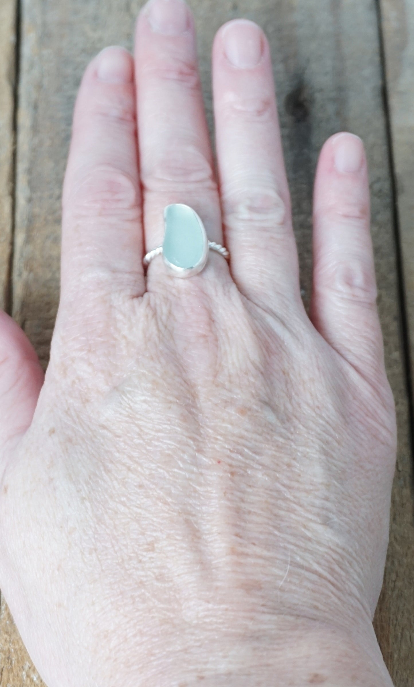 hand wearing a light aqua blue sea glass ring with a sterling silver setting, against a wooden background