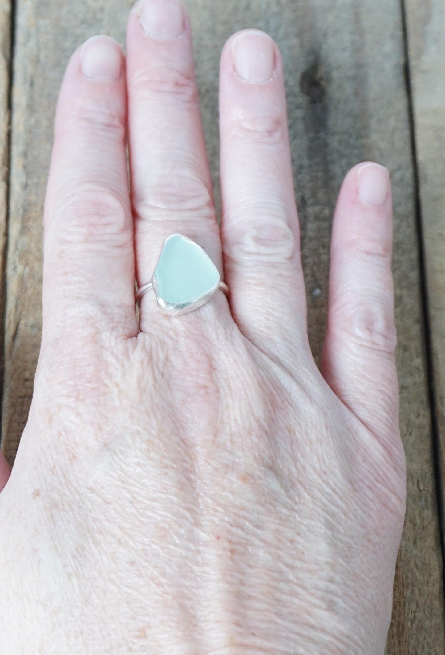 hand wearing a light aqua blue sea glass ring with a sterling silver setting, against a wooden background