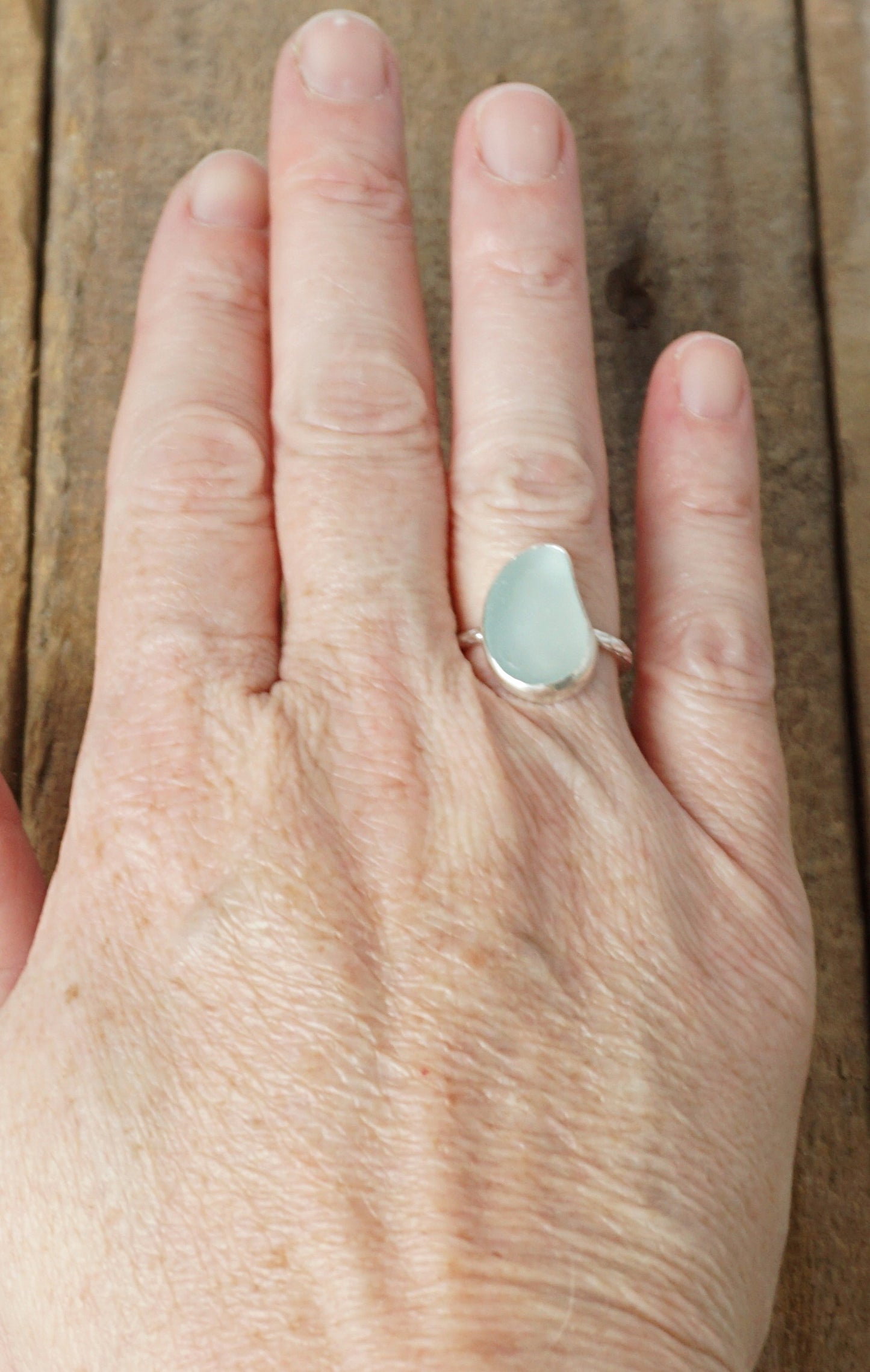 hand wearing a light aqua blue sea glass ring with a sterling silver setting, against a wooden background