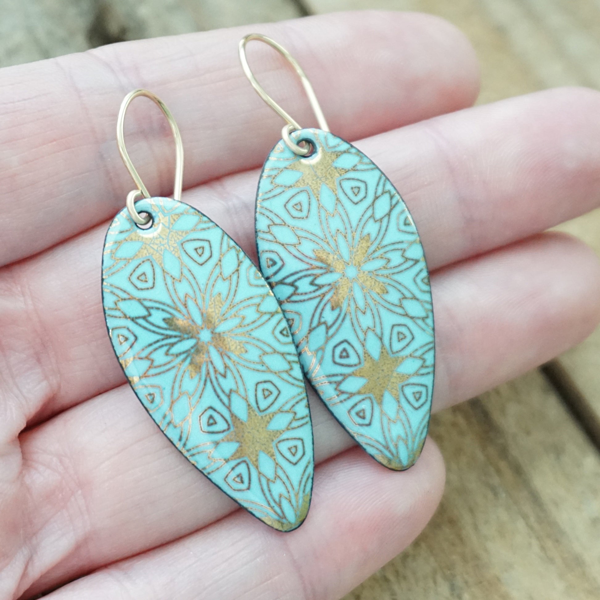 hand holding a pair of gold pattern on light blue enamel teardrop shaped earrings, on a wooden background