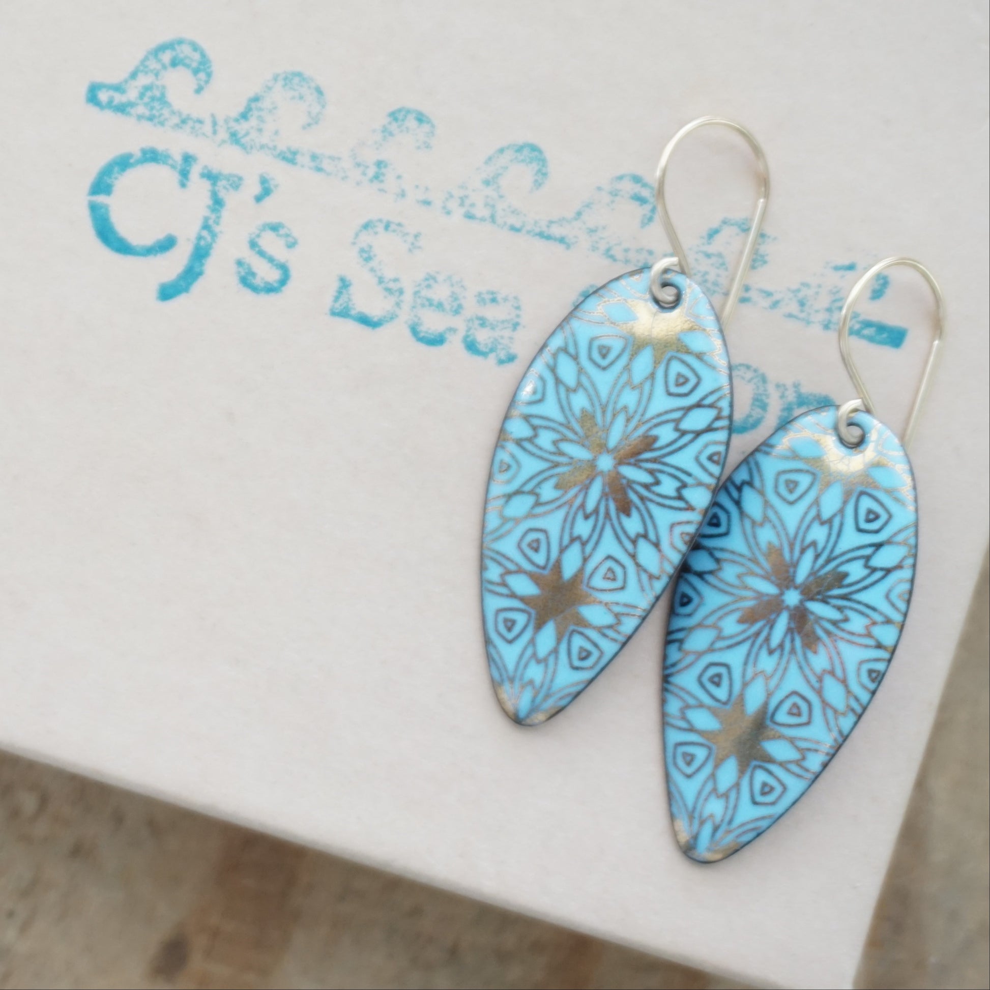 gold pattern on light blue enamel teardrop shaped earrings, on a gift box
