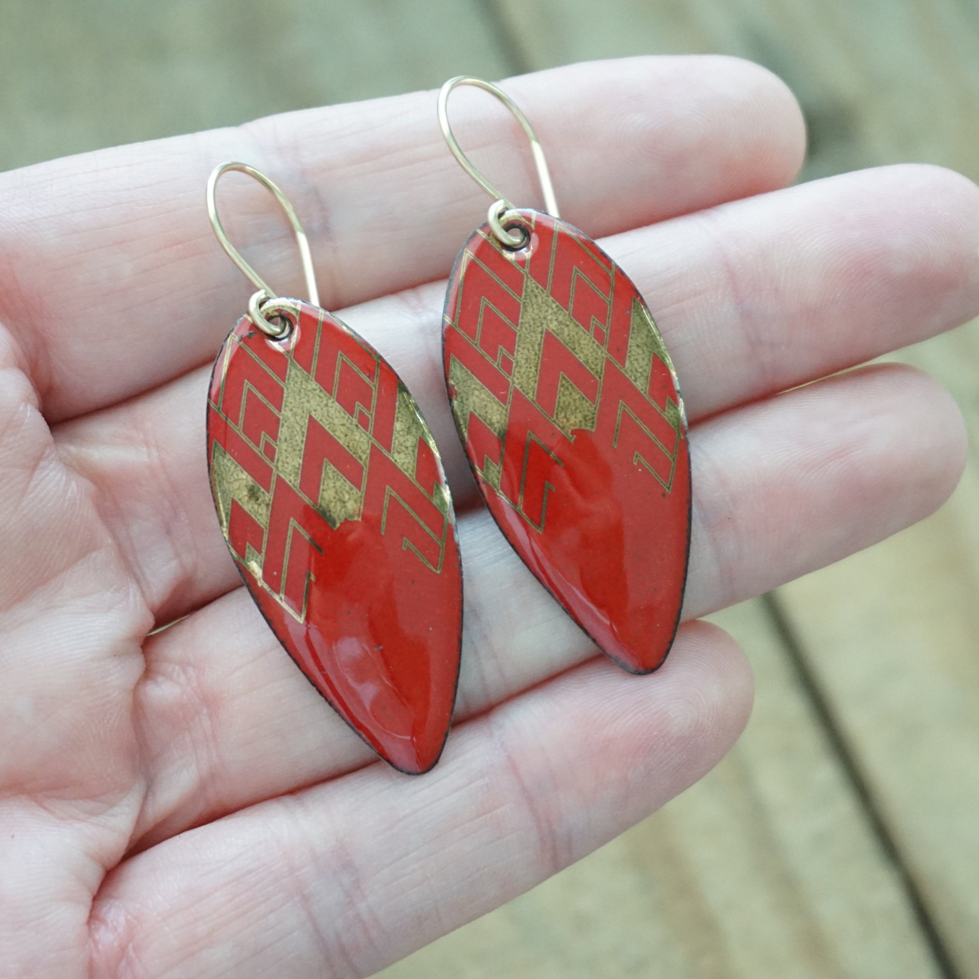 hand holding a pair of gold pattern on red enamel teardrop shaped earrings, on a wooden background