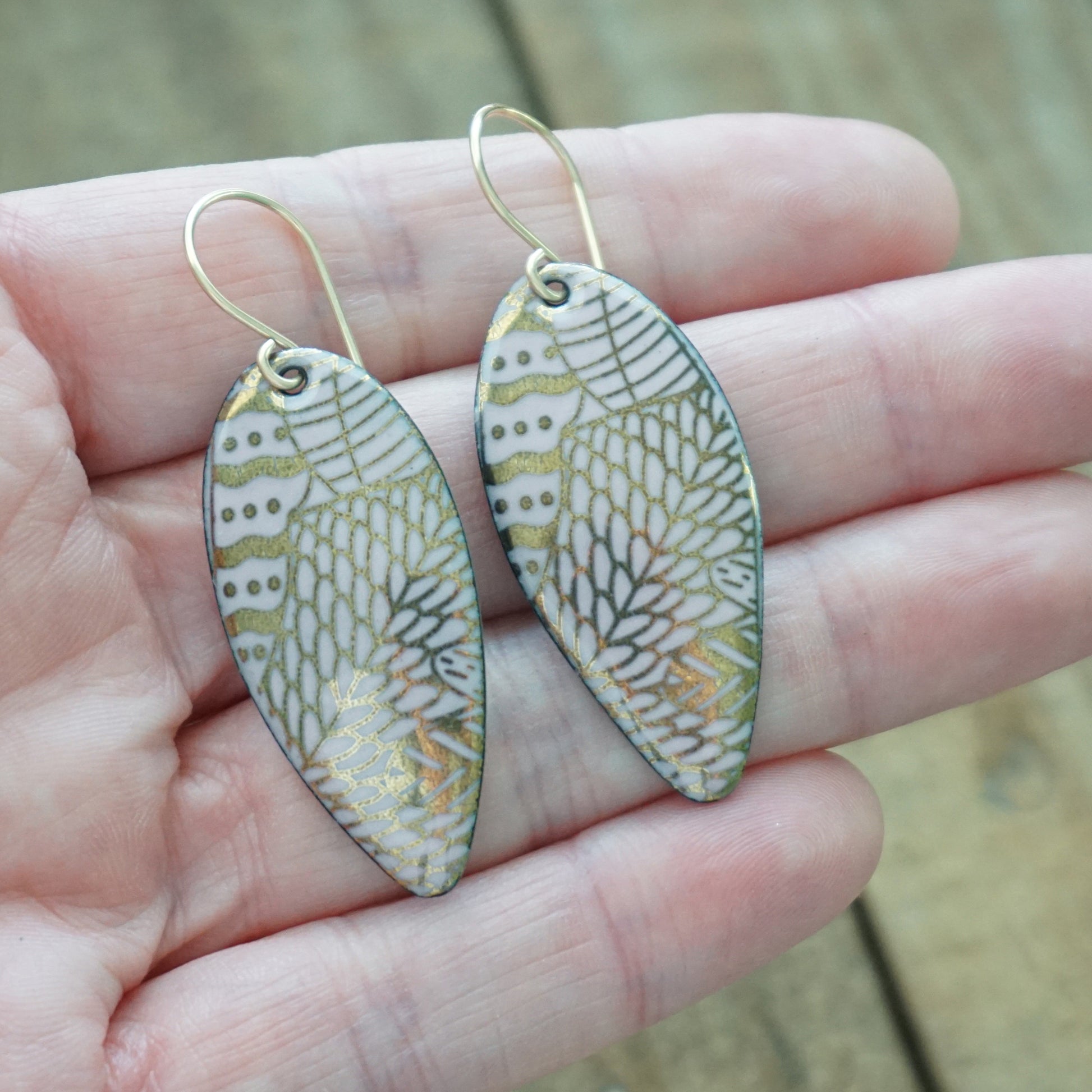 hand holding gold pattern on pink enamel teardrop shaped earrings, on a wooden background