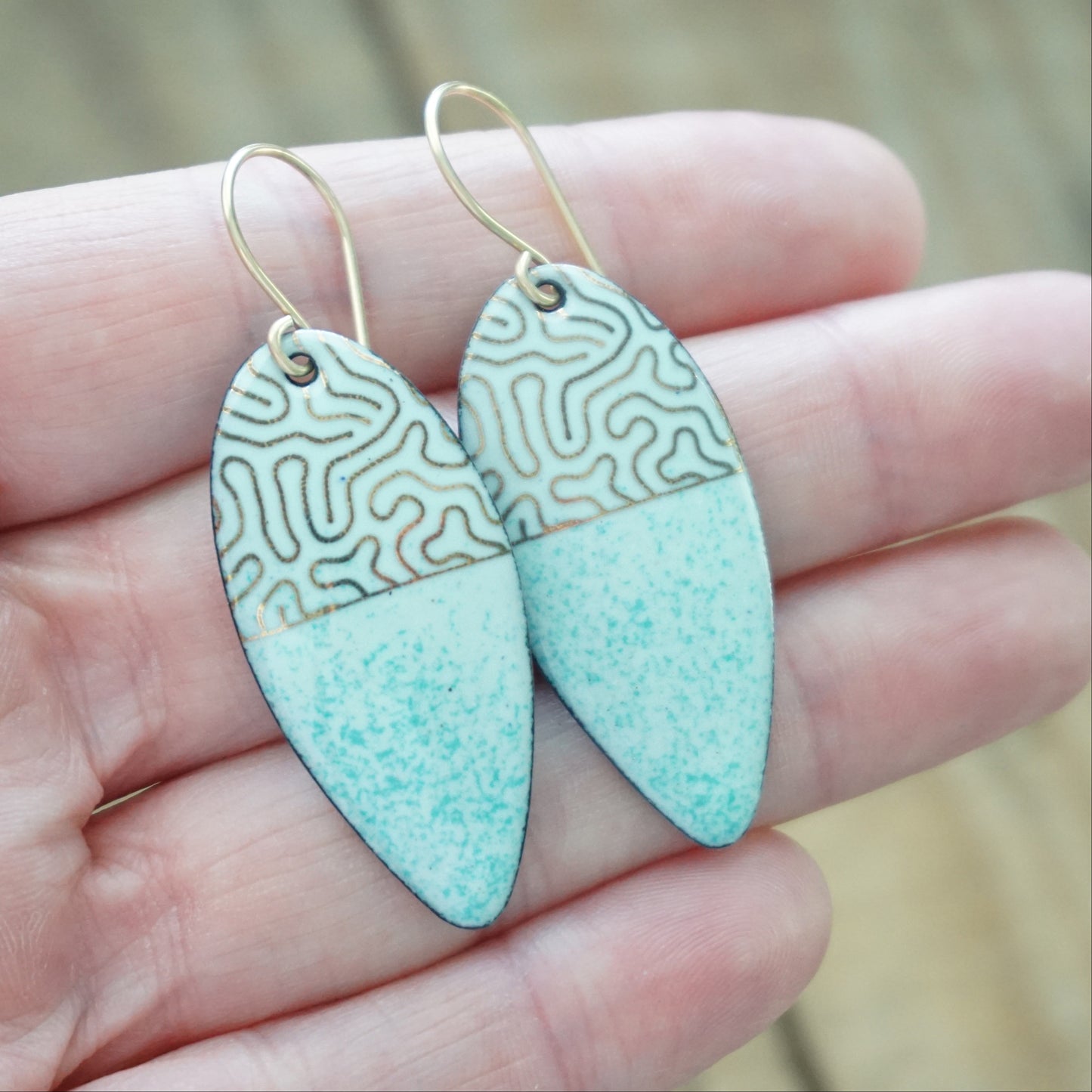 hand holding gold pattern on seafoam green enamel teardrop shaped earrings, on a wooden background