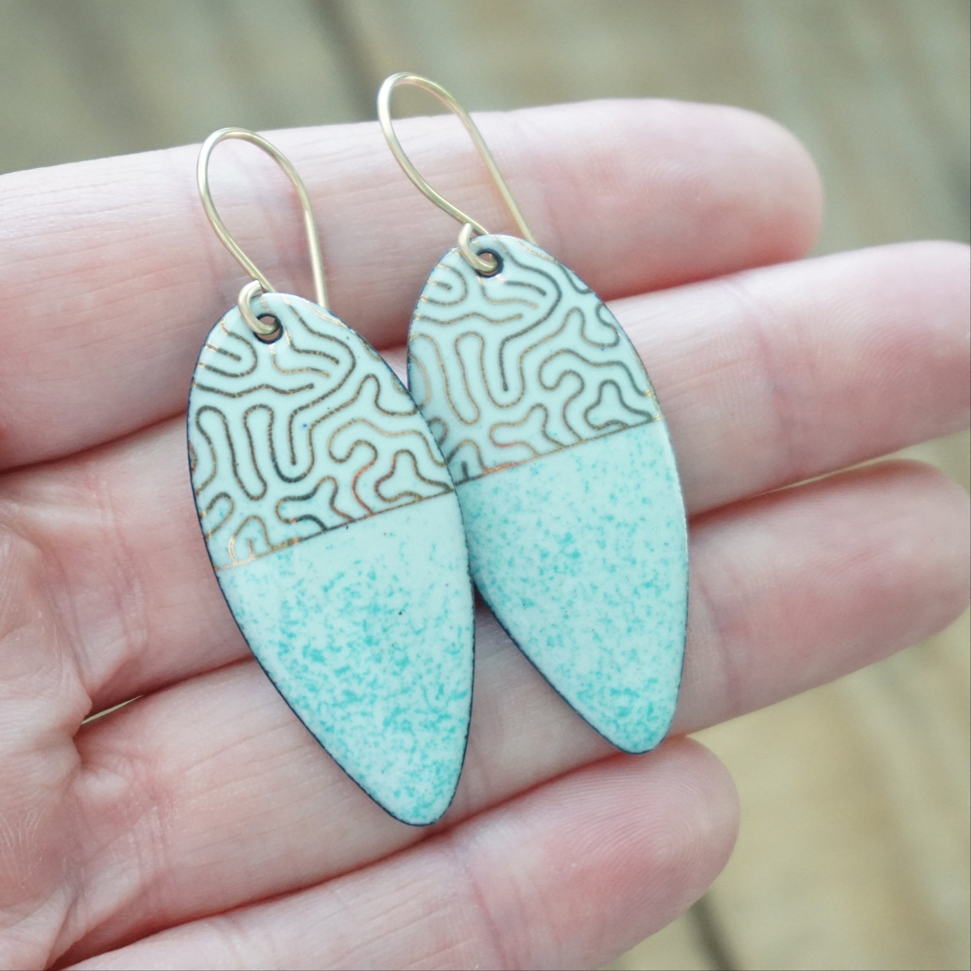 hand holding gold pattern on seafoam green enamel teardrop shaped earrings, on a wooden background
