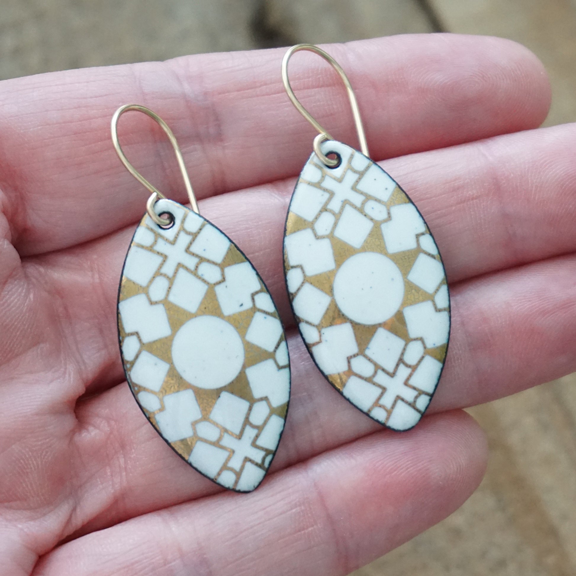 hand holding a pair of White, marquis shaped earrings with gold accents, against a wooden background