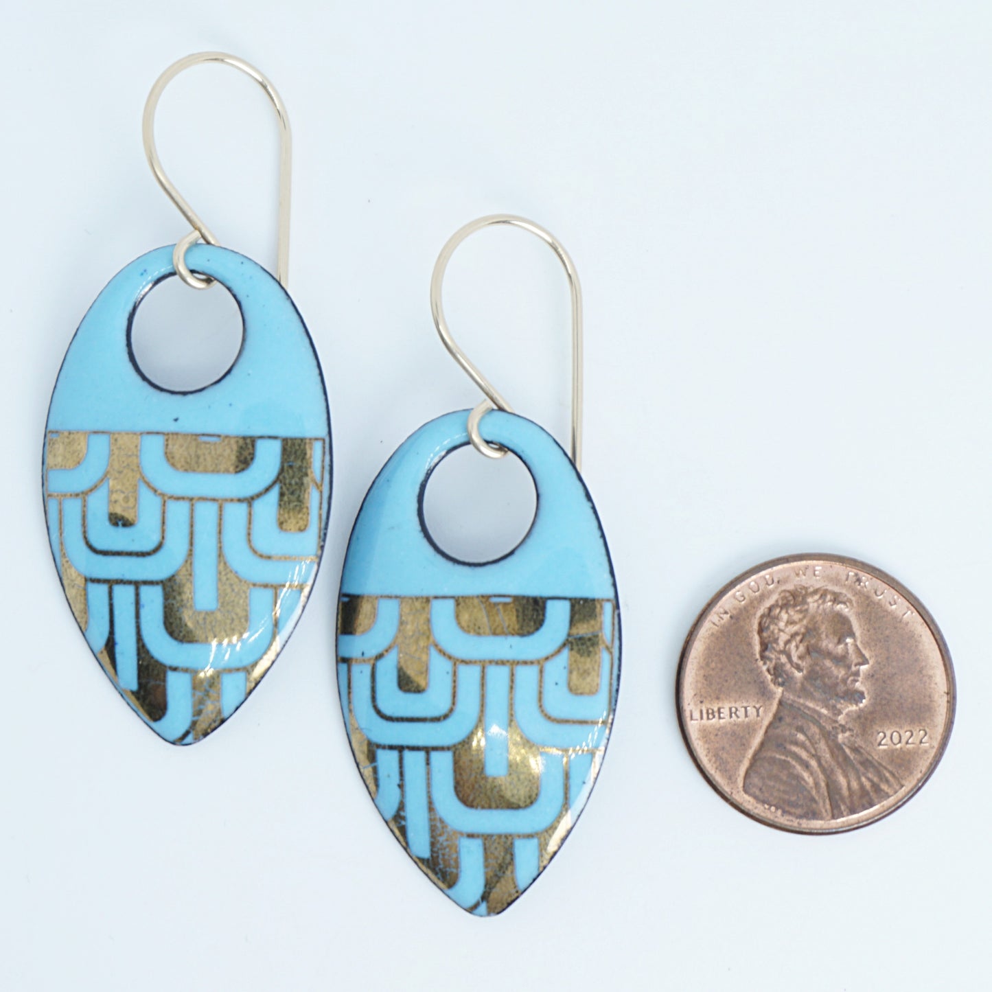 Light blue, teardrop shaped earrings with gold accents, next to a penny for scale, against a white background