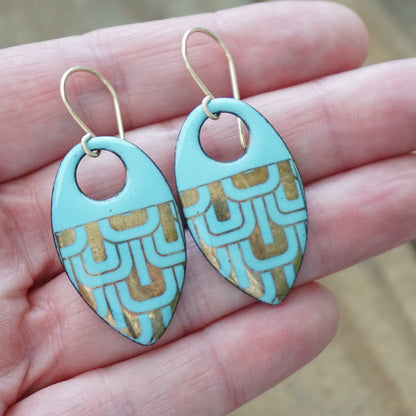 Hand holding a pair of Light blue, teardrop shaped earrings with gold accents, against a wooden background