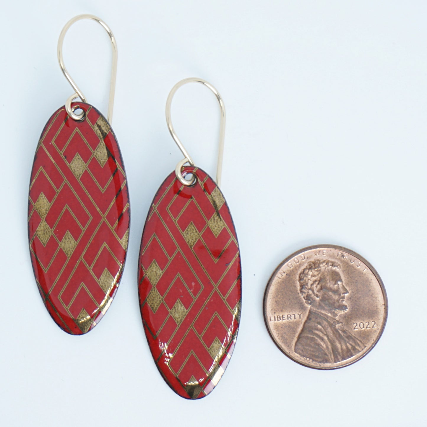 Red, oval shaped earrings with gold accents, next to a penny for scale, against a white background