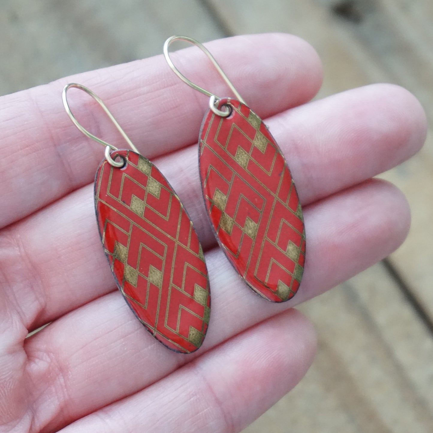 Hand holding a pair of Red, oval shaped earrings with gold accents, against a wooden background