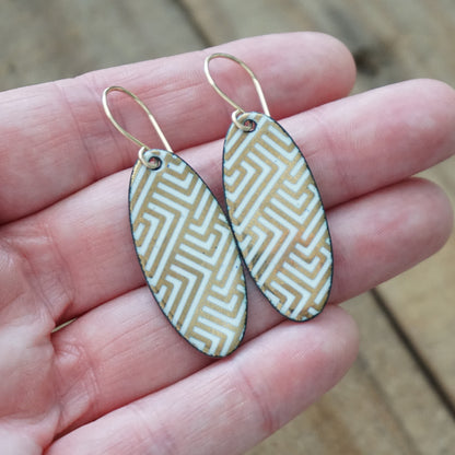 Hand holding a pair of White, oval shaped earrings with gold accents, against a wooden background