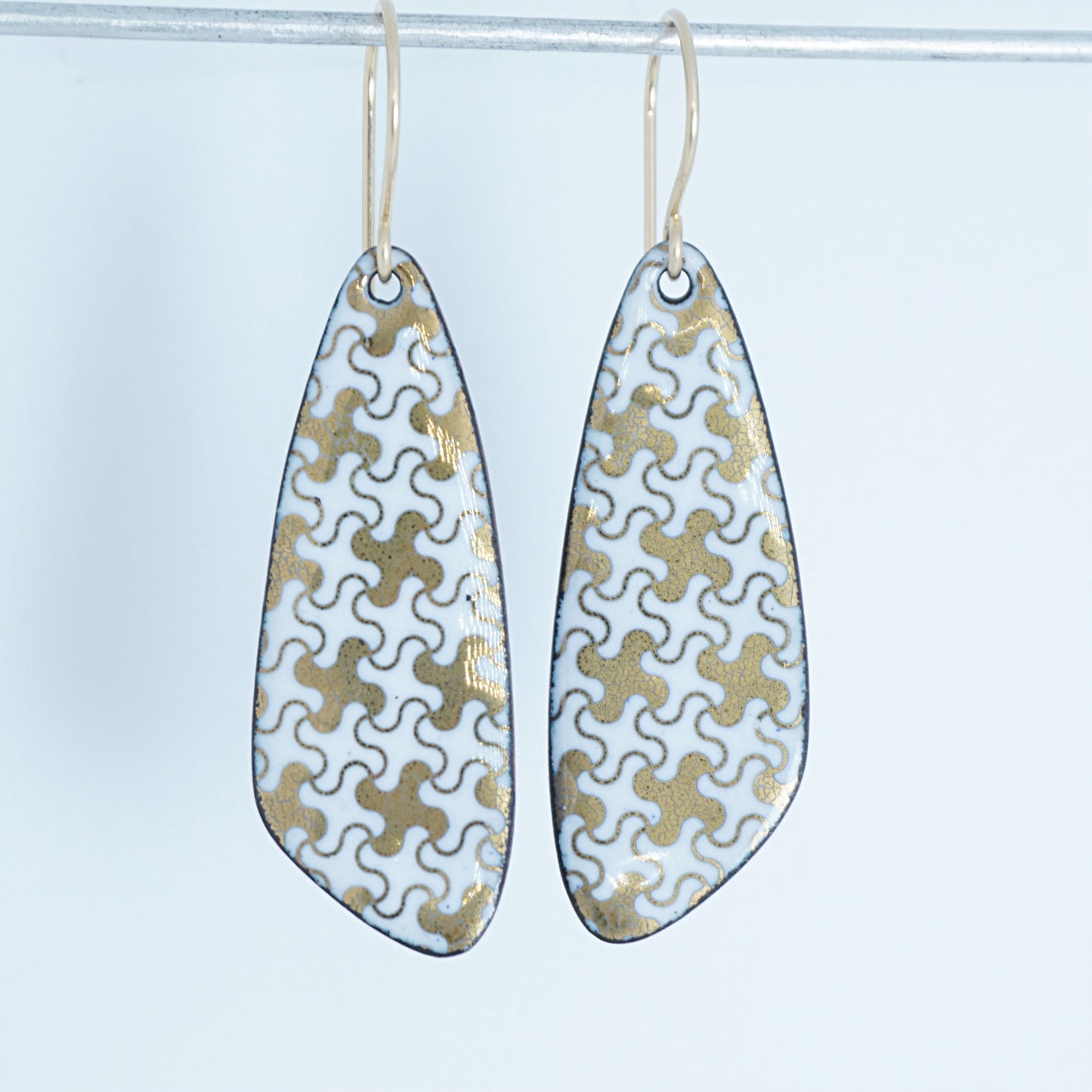 A pair of teardrop-shaped dangle earrings with a white enamel background, decorated with a gold puzzle-piece pattern and hanging from thin gold hooks.