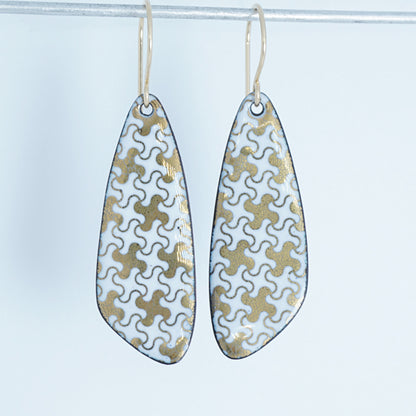 A pair of teardrop-shaped dangle earrings with a white enamel background, decorated with a gold puzzle-piece pattern and hanging from thin gold hooks.