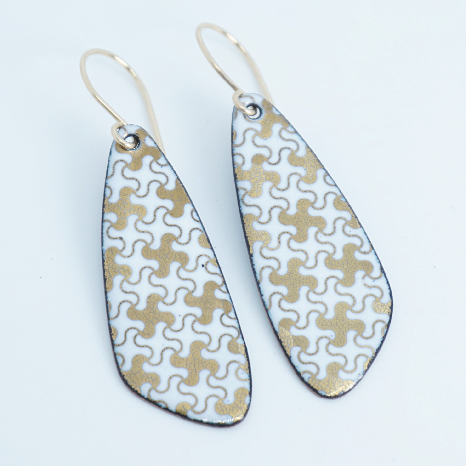 A pair of teardrop-shaped dangle earrings with a white and gold interlocking puzzle piece pattern, suspended from thin gold fishhook wires, shown on a plain white background.