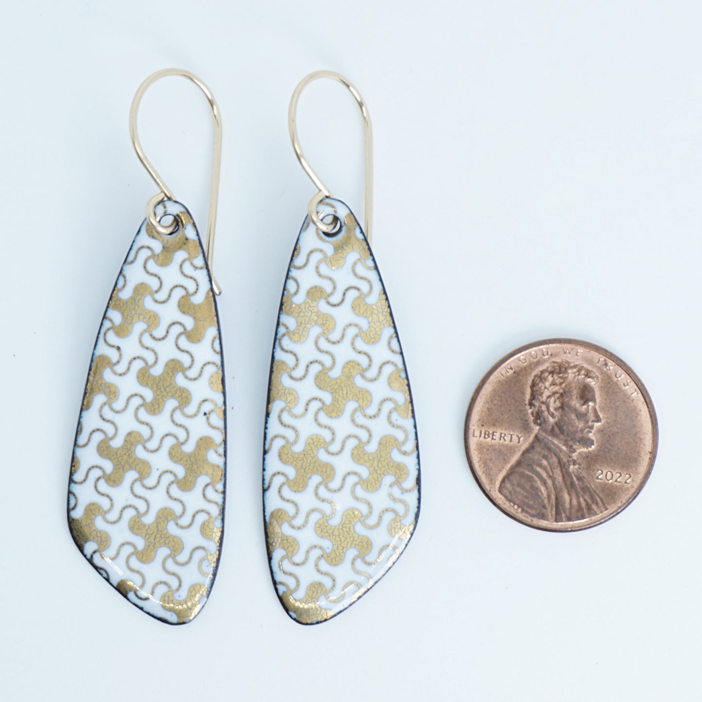 A pair of teardrop-shaped drop earrings with a white and gold puzzle-piece pattern and gold fishhook wires, shown next to a penny for scale on a white background.