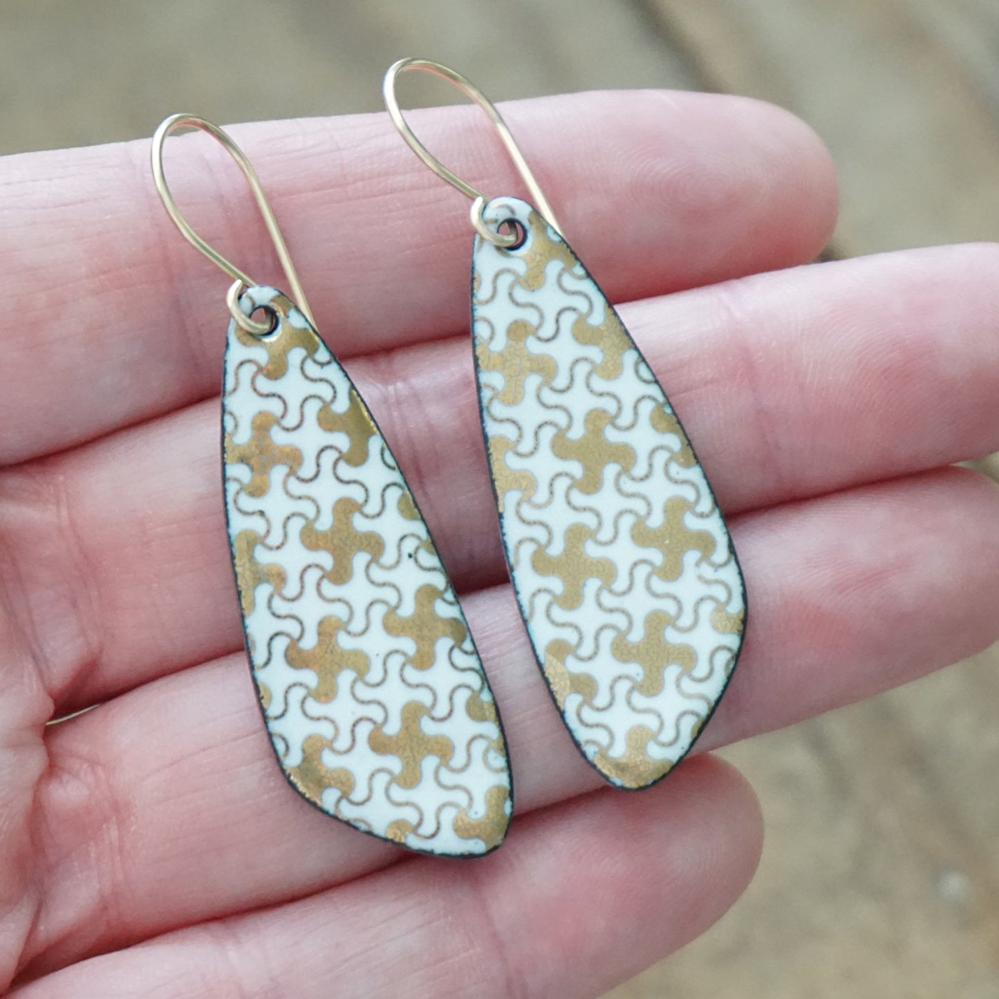 A pair of teardrop-shaped drop earrings with a gold and white puzzle-piece pattern, held in a person's hand.