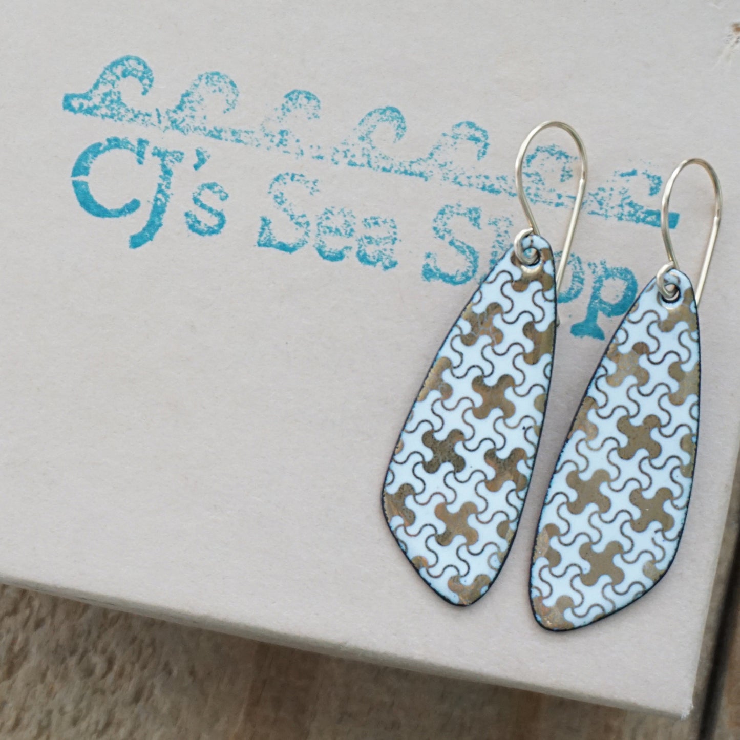 A pair of dangling teardrop earrings with a light blue and tan puzzle-piece pattern, displayed on a box with a blue 'CJ's Sea Shop' logo.