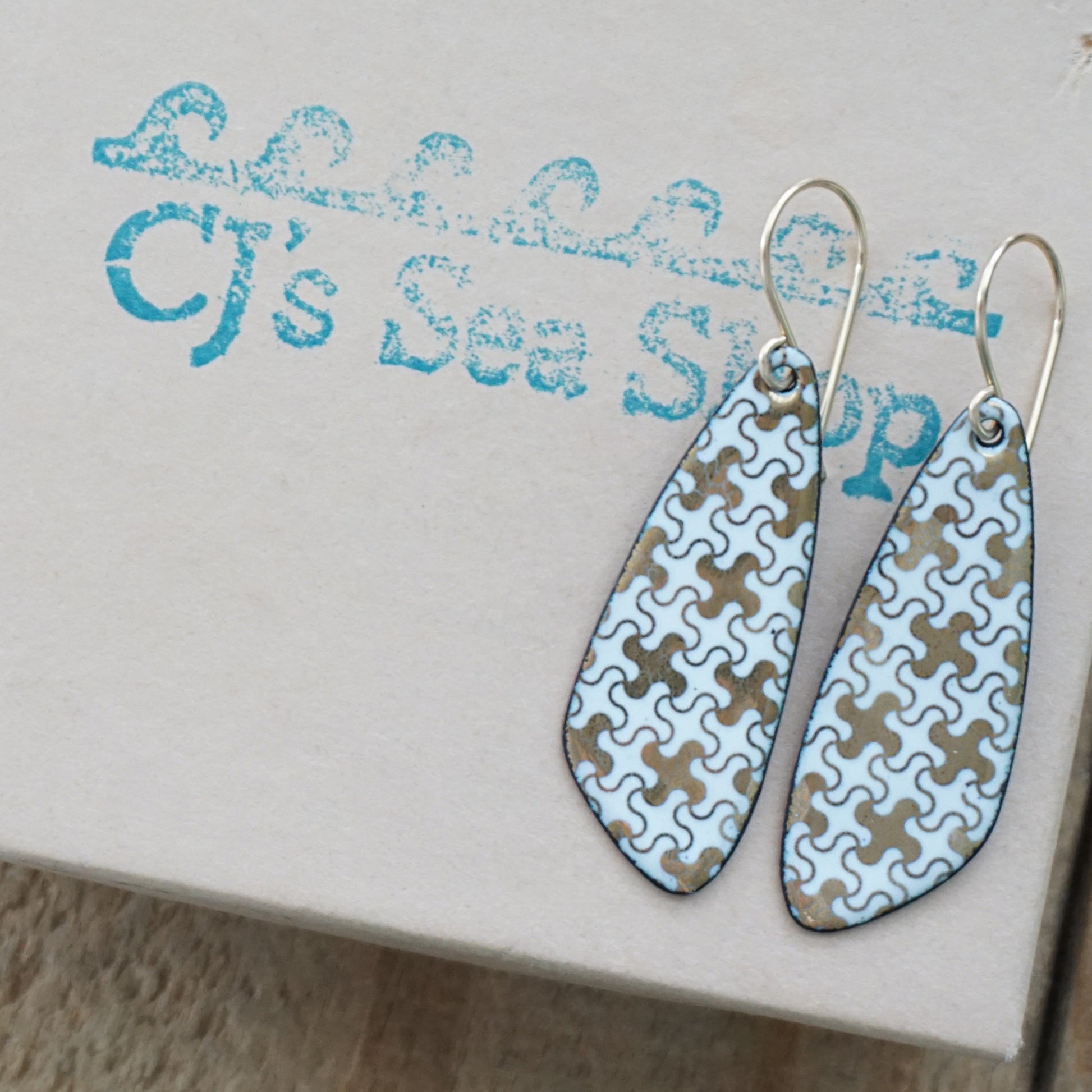 A pair of dangling teardrop earrings with a light blue and tan puzzle-piece pattern, displayed on a box with a blue 'CJ's Sea Shop' logo.