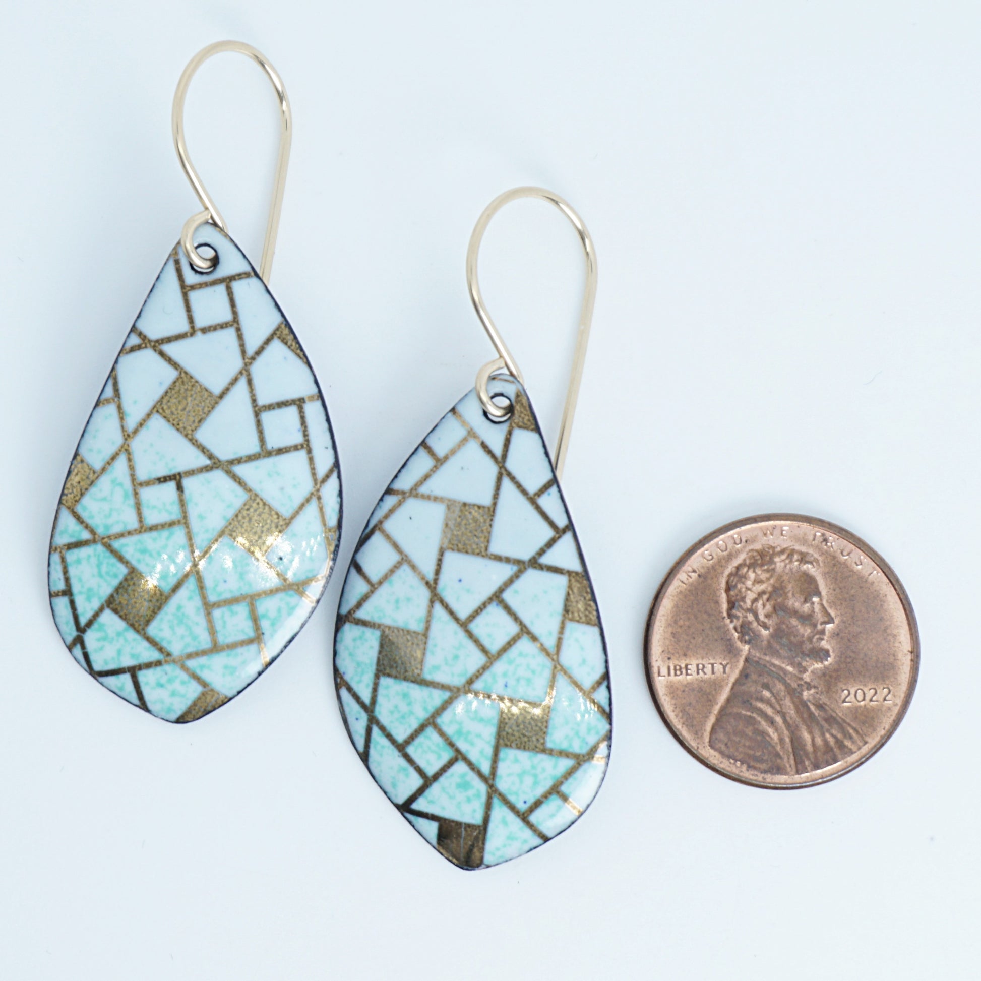 A pair of leaf-shaped dangling earrings with a mosaic pattern of light green and gold pieces, shown next to a penny for scale on a white background