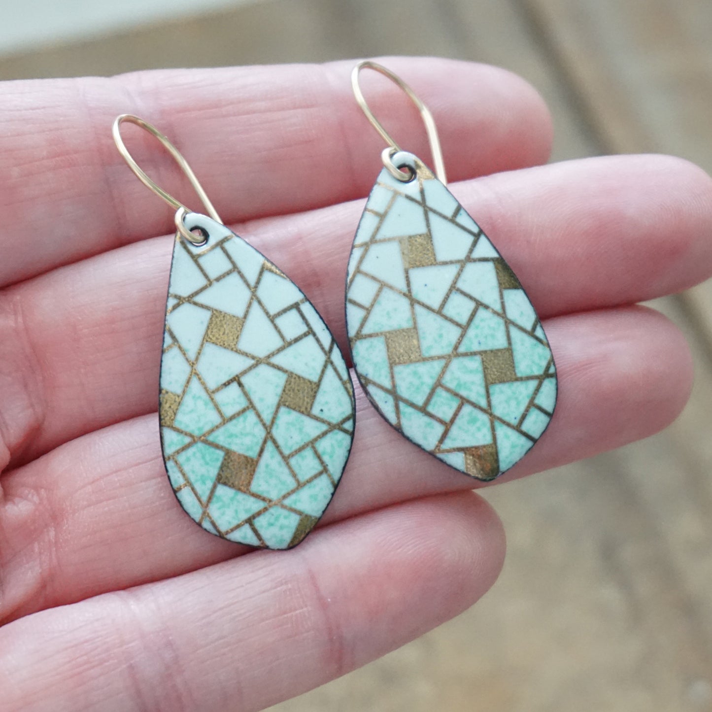A pair of leaf-shaped dangling earrings with a mosaic pattern of light  green and gold geometric shapes, held in a person's hand.