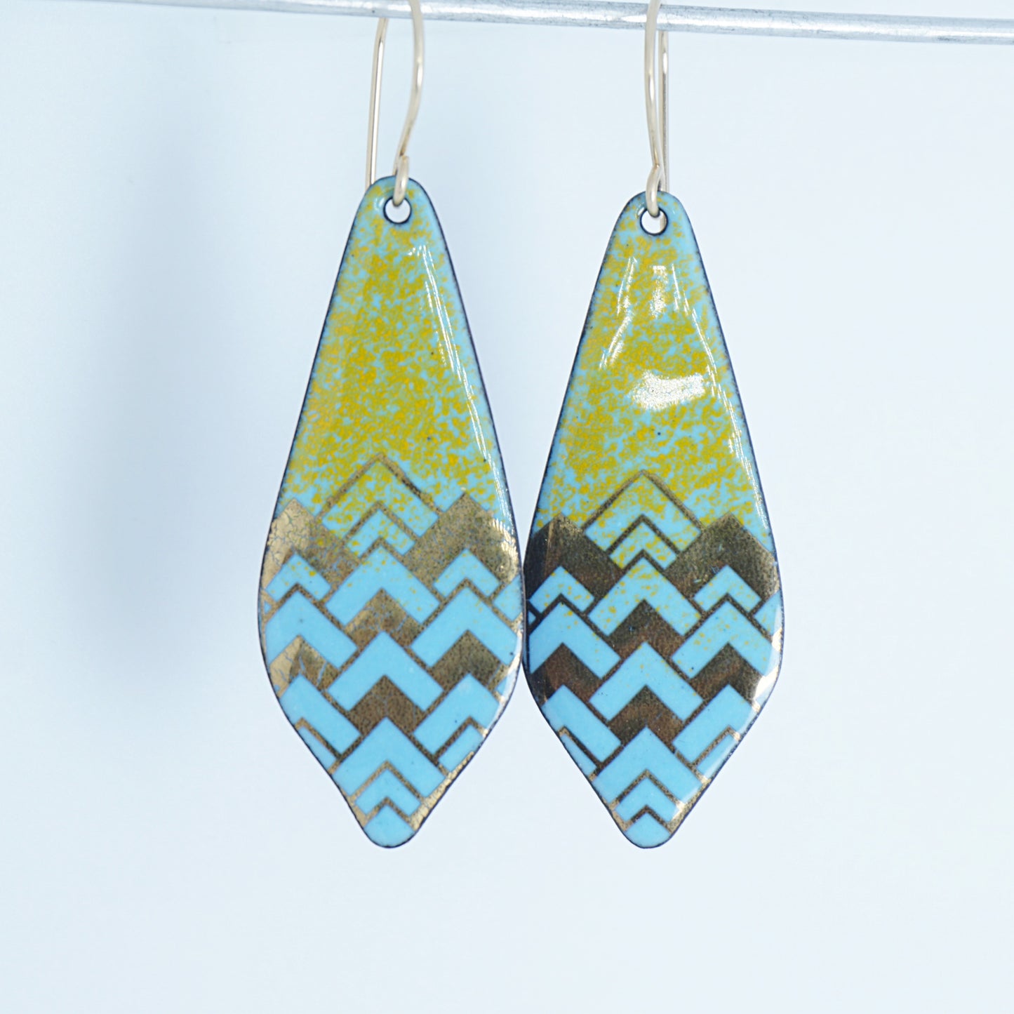 A pair of dangling teardrop-shaped earrings with a colorful geometric pattern. The top half of each earring is orange, and the bottom half features a chevron pattern in light blue and gold, against a white background