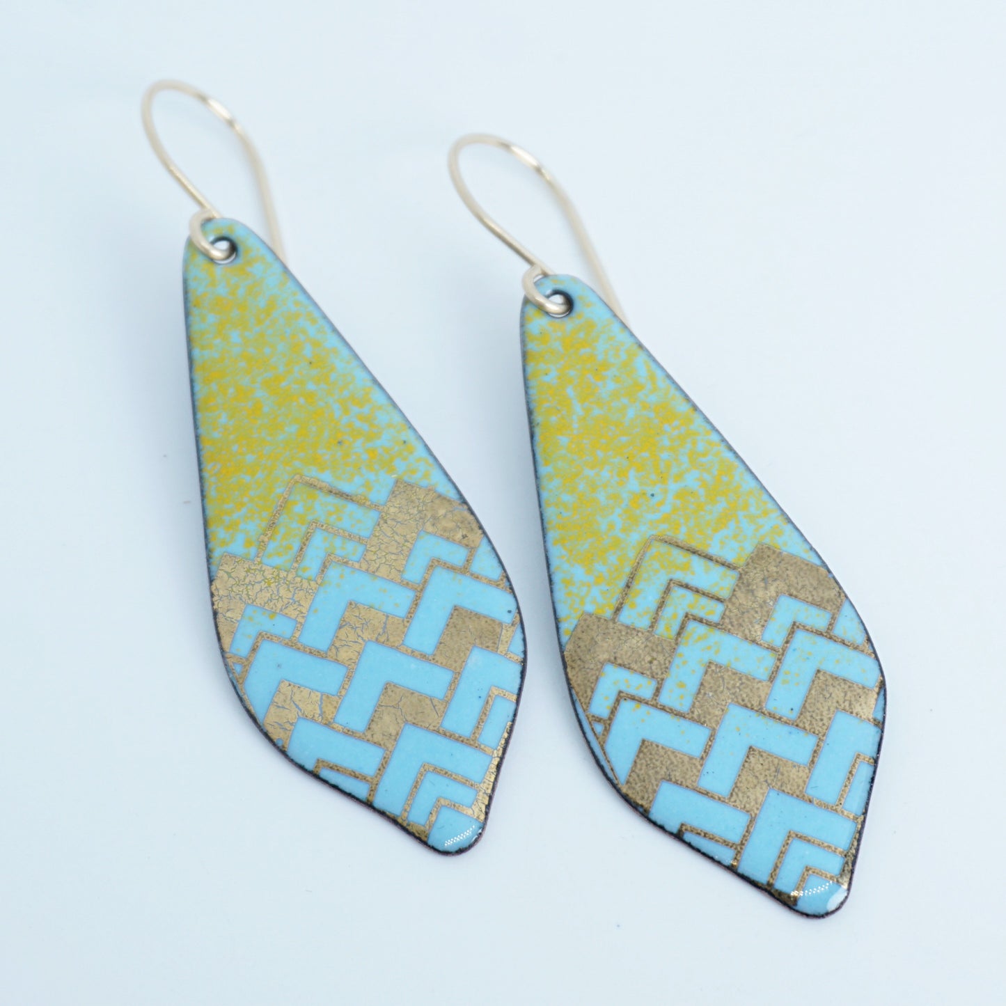 A pair of dangling teardrop-shaped earrings with a colorful geometric pattern. The top half of each earring is orange, and the bottom half features a chevron pattern in light blue and gold, against a white background