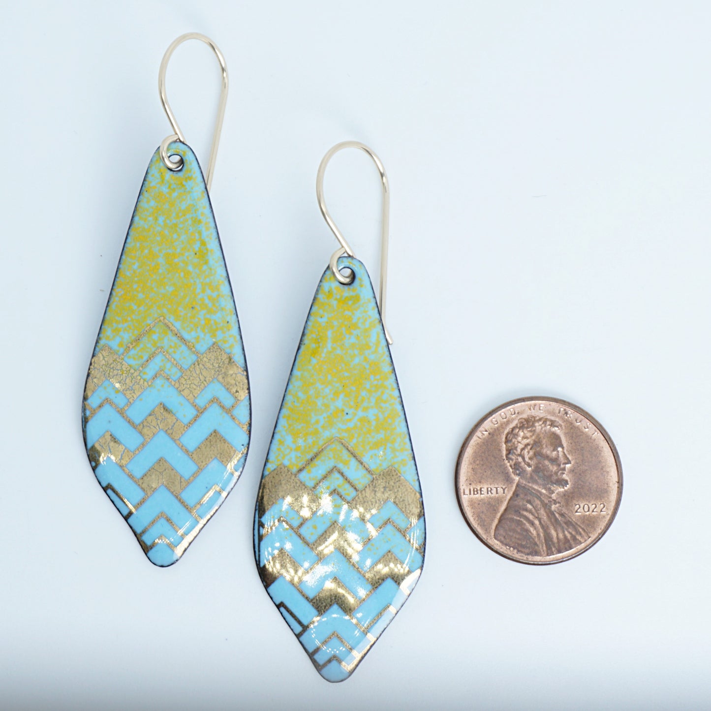 A pair of dangling teardrop-shaped earrings with a colorful geometric pattern. The top half of each earring is orange, and the bottom half features a chevron pattern in light blue and gold, next to a penny for scale, against a white background