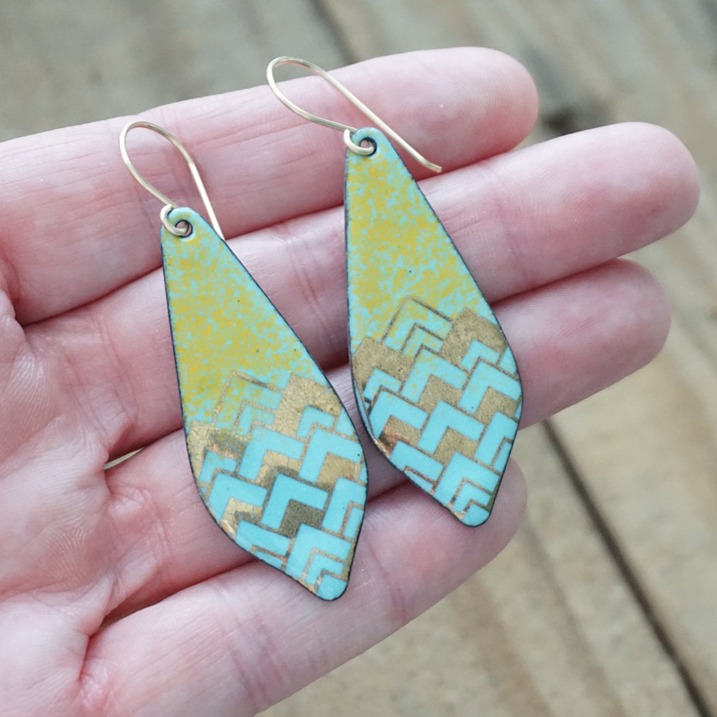 A pair of teardrop-shaped drop earrings with a gold-tone wire hook and a light blue, orange, and gold chevron pattern, held in a person's hand against a blurred wooden background.