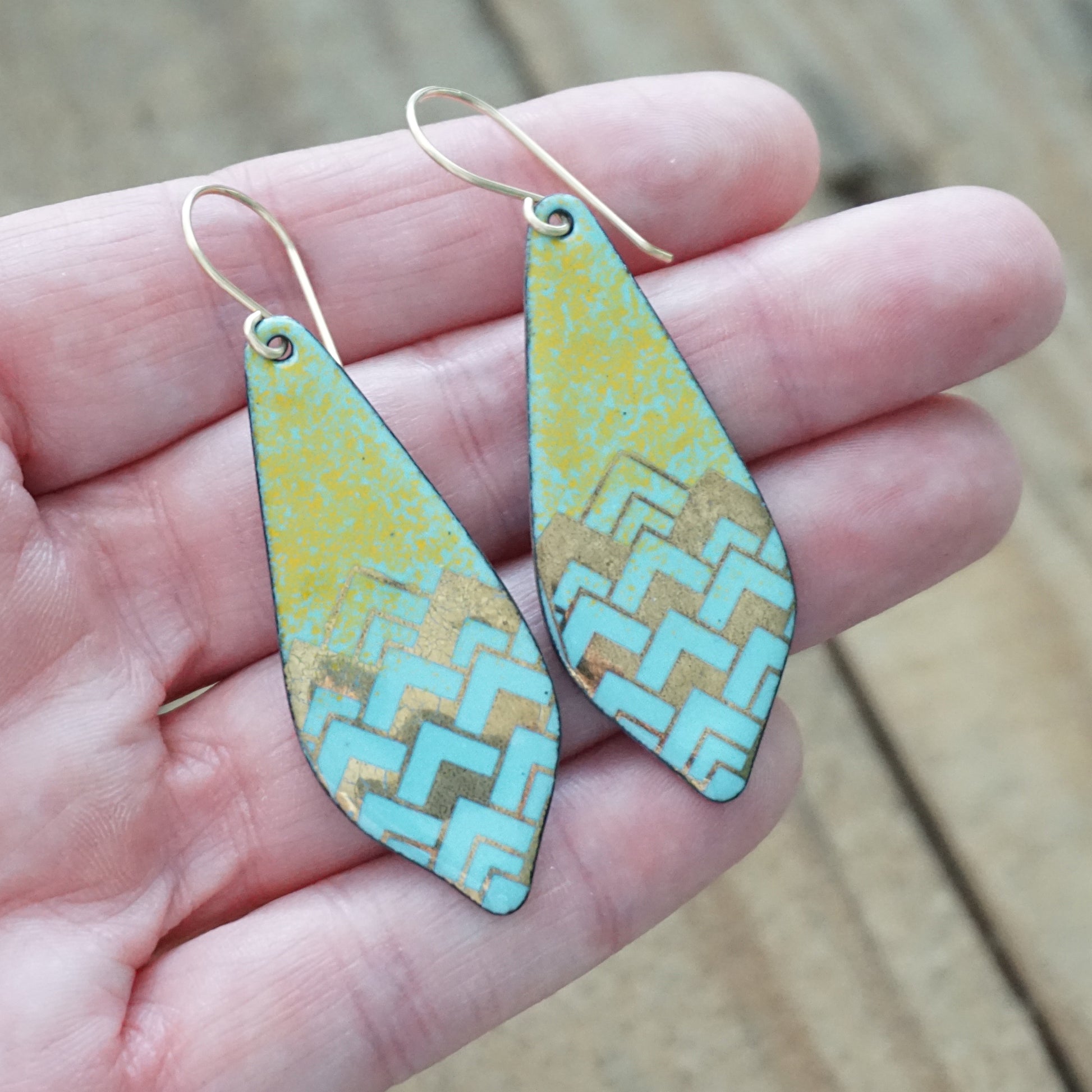 A pair of teardrop-shaped drop earrings with a gold-tone wire hook and a light blue, orange, and gold chevron pattern, held in a person's hand against a blurred wooden background.