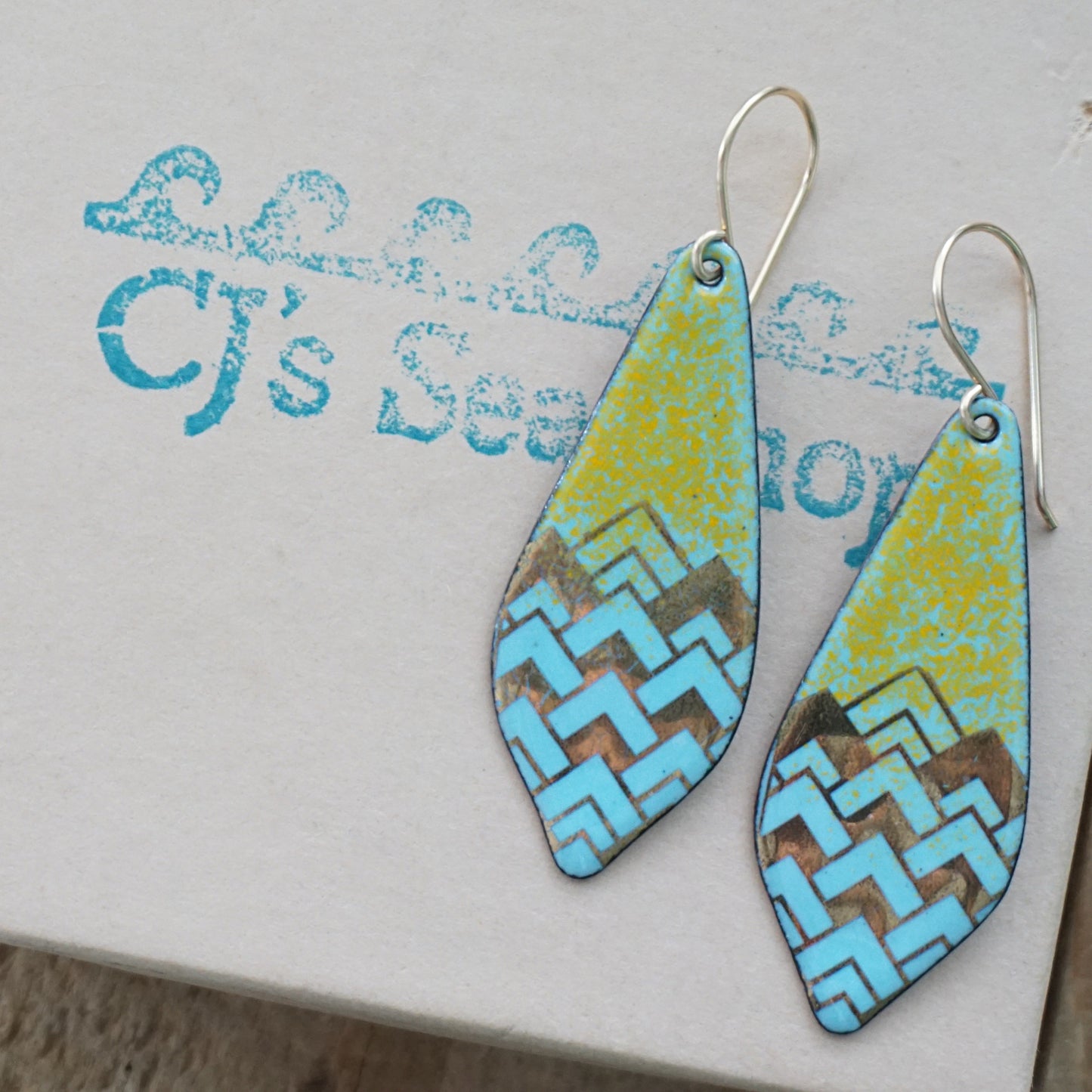 A pair of dangling teardrop-shaped earrings with a colorful geometric pattern. The top half of each earring is orange, and the bottom half features a chevron pattern in light blue and gold, on a gift box