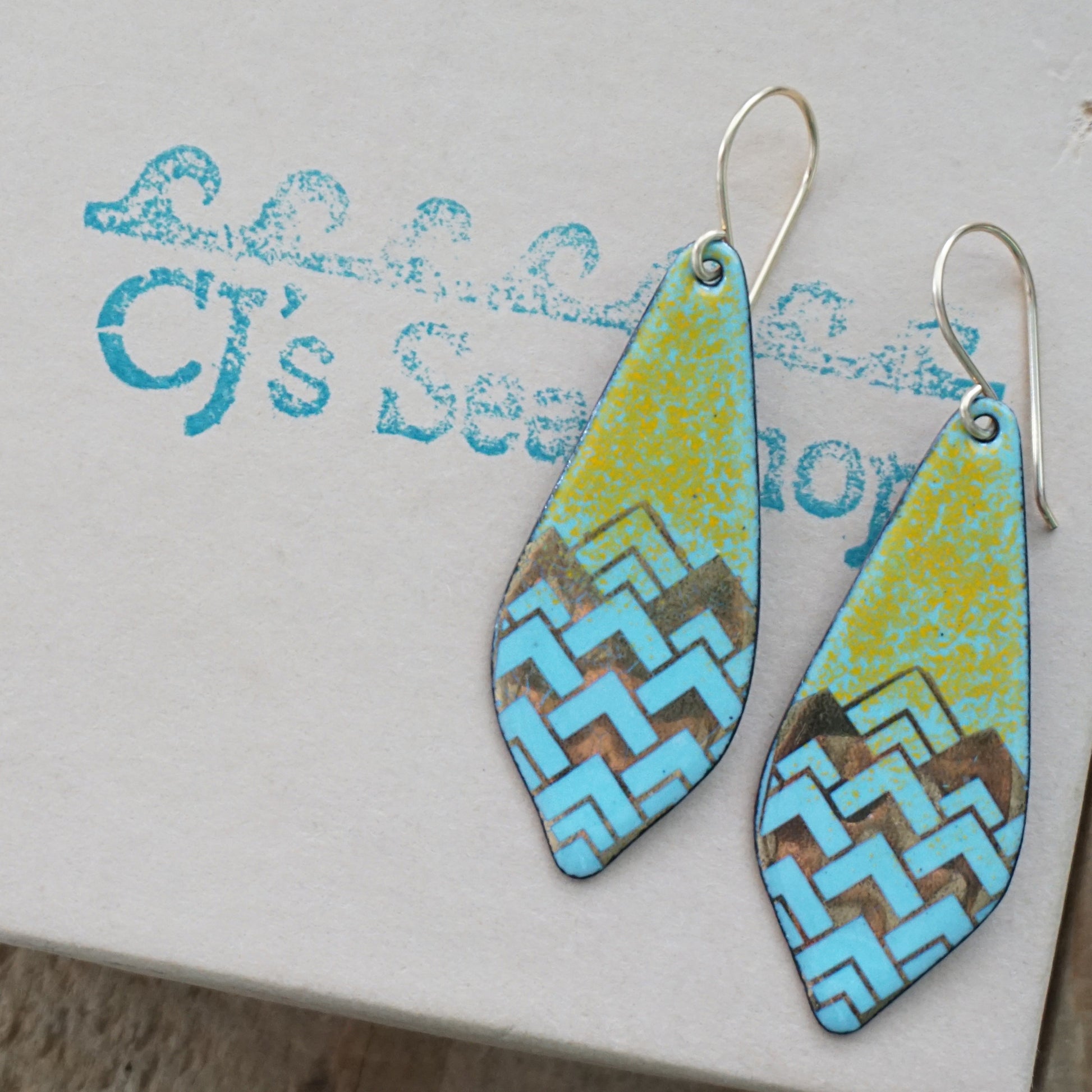 A pair of dangling teardrop-shaped earrings with a colorful geometric pattern. The top half of each earring is orange, and the bottom half features a chevron pattern in light blue and gold, on a gift box