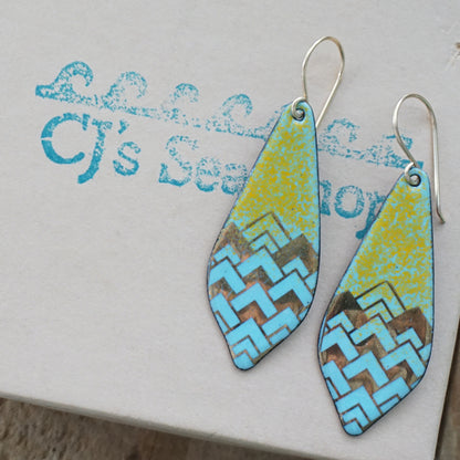 A pair of dangling teardrop-shaped earrings with a colorful geometric pattern. The top half of each earring is orange, and the bottom half features a chevron pattern in light blue and gold, on a gift box