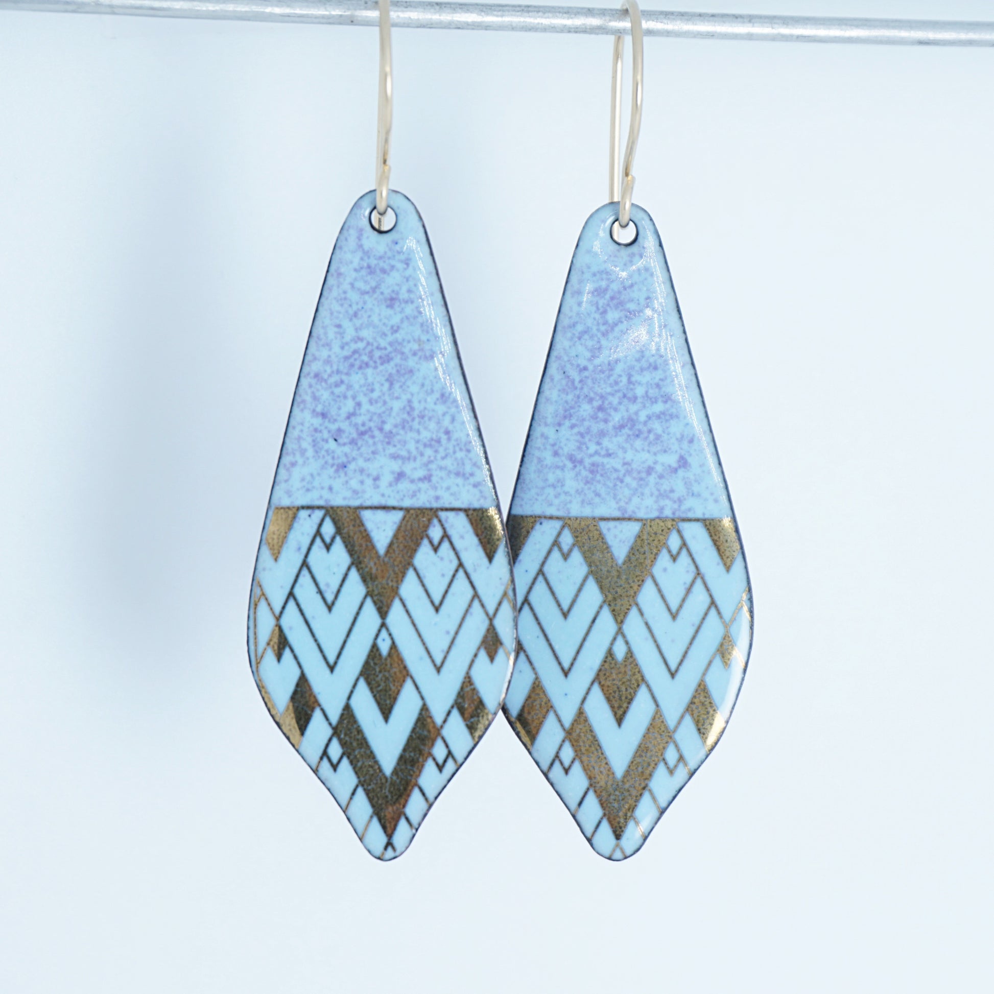 A pair of teardrop-shaped dangle earrings with a light blue and purple top half and a geometric chevron pattern in light blue and metallic gold on the bottom half, hanging from thin gold hooks against a plain light background.