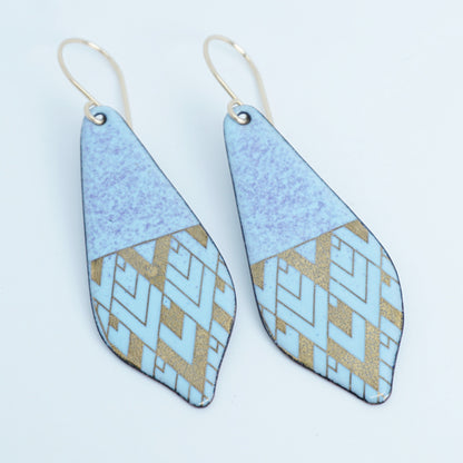 A pair of dangling geometric earrings with a light blue and purple top half and a lower half featuring a light blue and gold geometric pattern, suspended from gold fishhook wires on a white background.