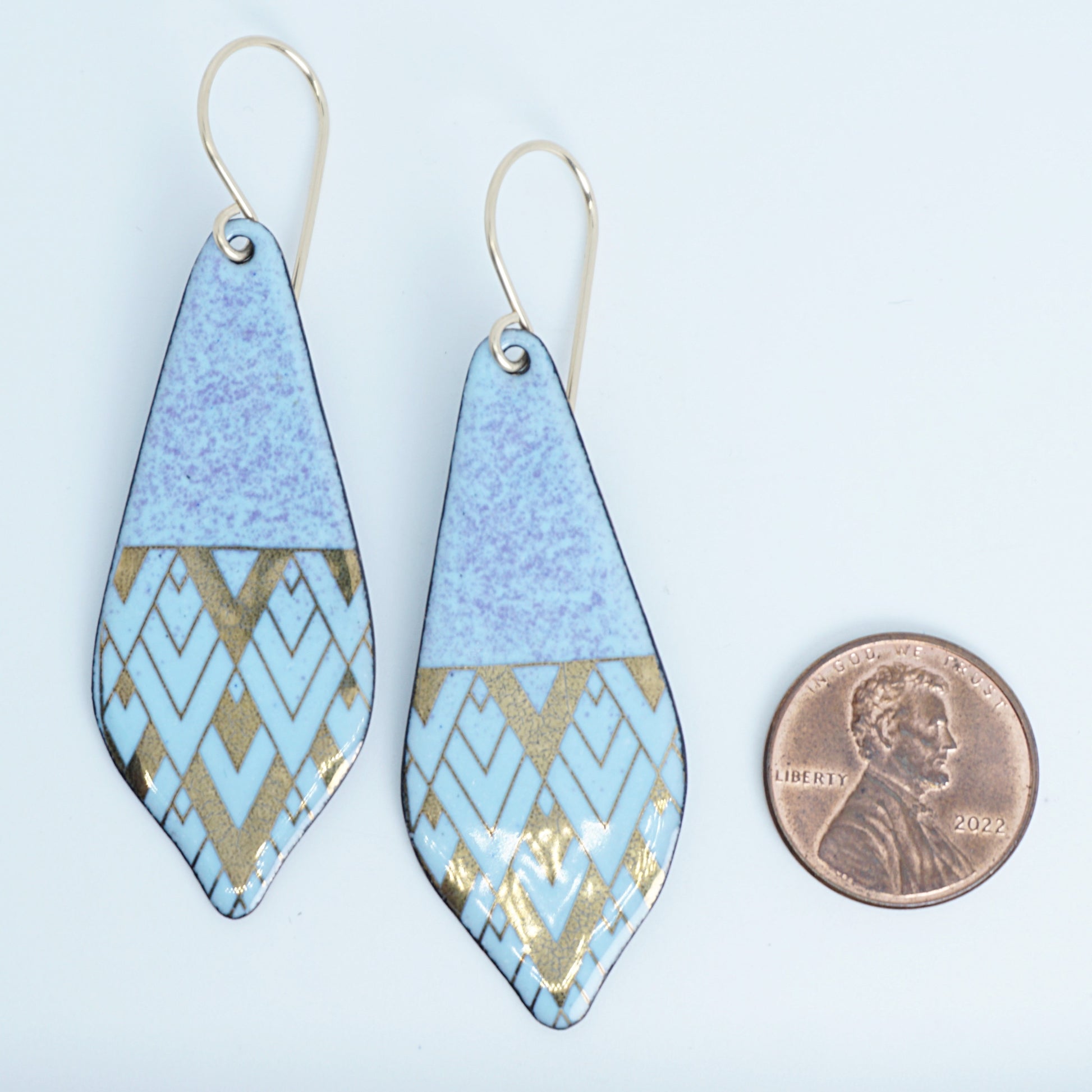 A pair of dangling teardrop earrings with a light blue, purple, and gold geometric pattern and gold-tone metal hooks, shown next to a penny for scale on a white background.