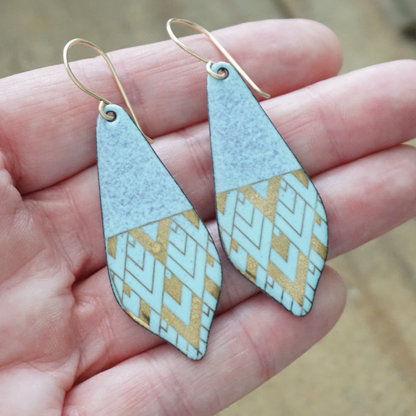 A pair of handmade teardrop-shaped dangle earrings held in a person's hand. The earrings feature a light blue and purple top half and a lower half with a gold geometric pattern on a light blue background, suspended from gold fishhook wires.
