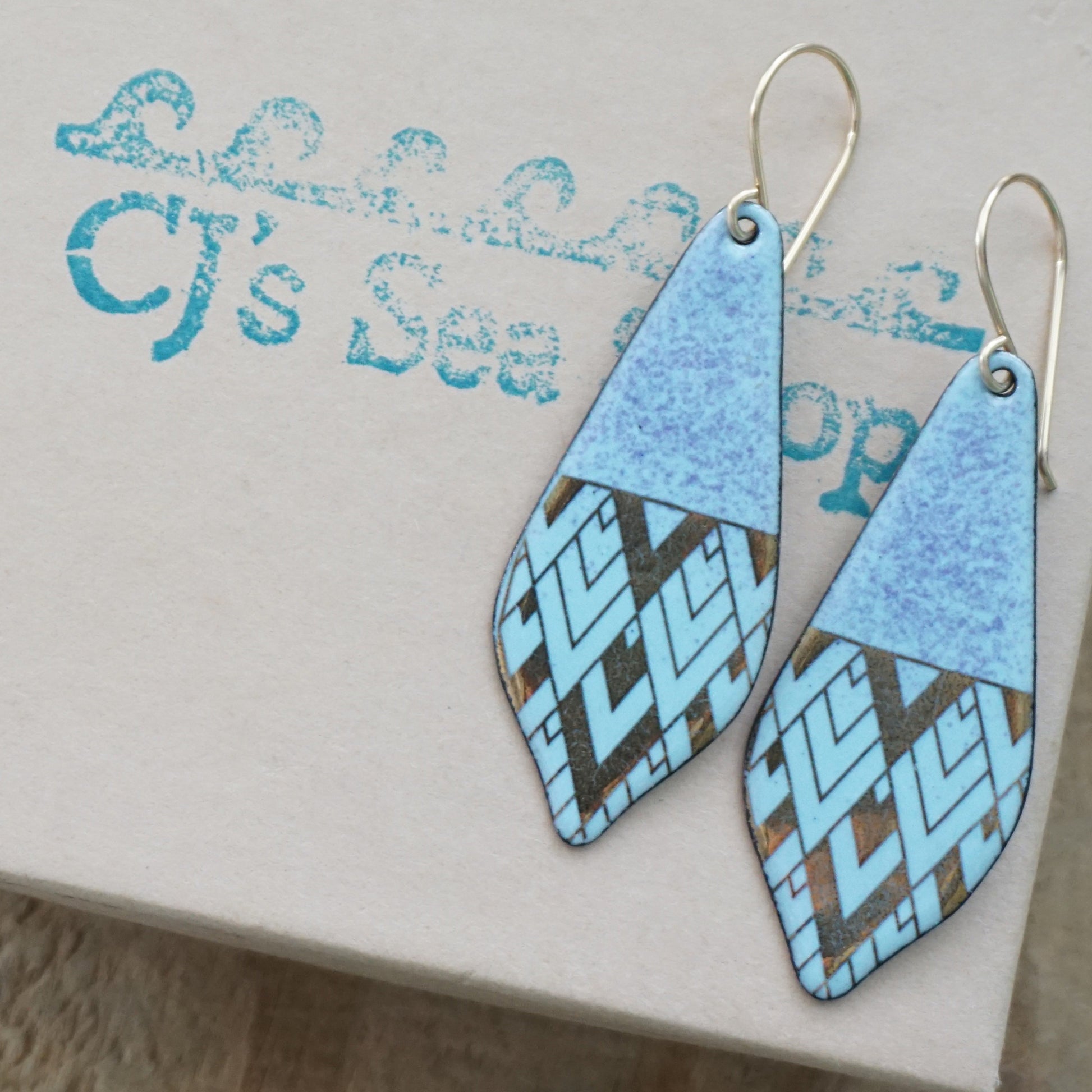 A pair of handmade teardrop-shaped dangle earrings with a light blue and purple top section and a gold and light blue geometric patterned bottom section, displayed on a gift box.