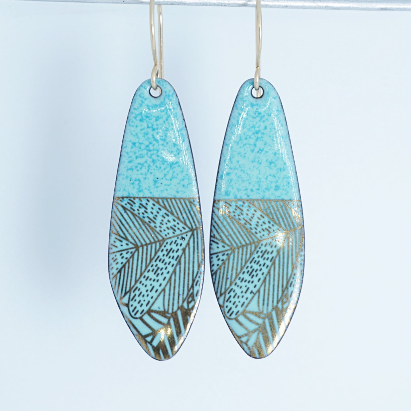 A pair of seafoam green teardrop shaped earrings with gold abstract accents, suspended from thin gold hooks on a white background