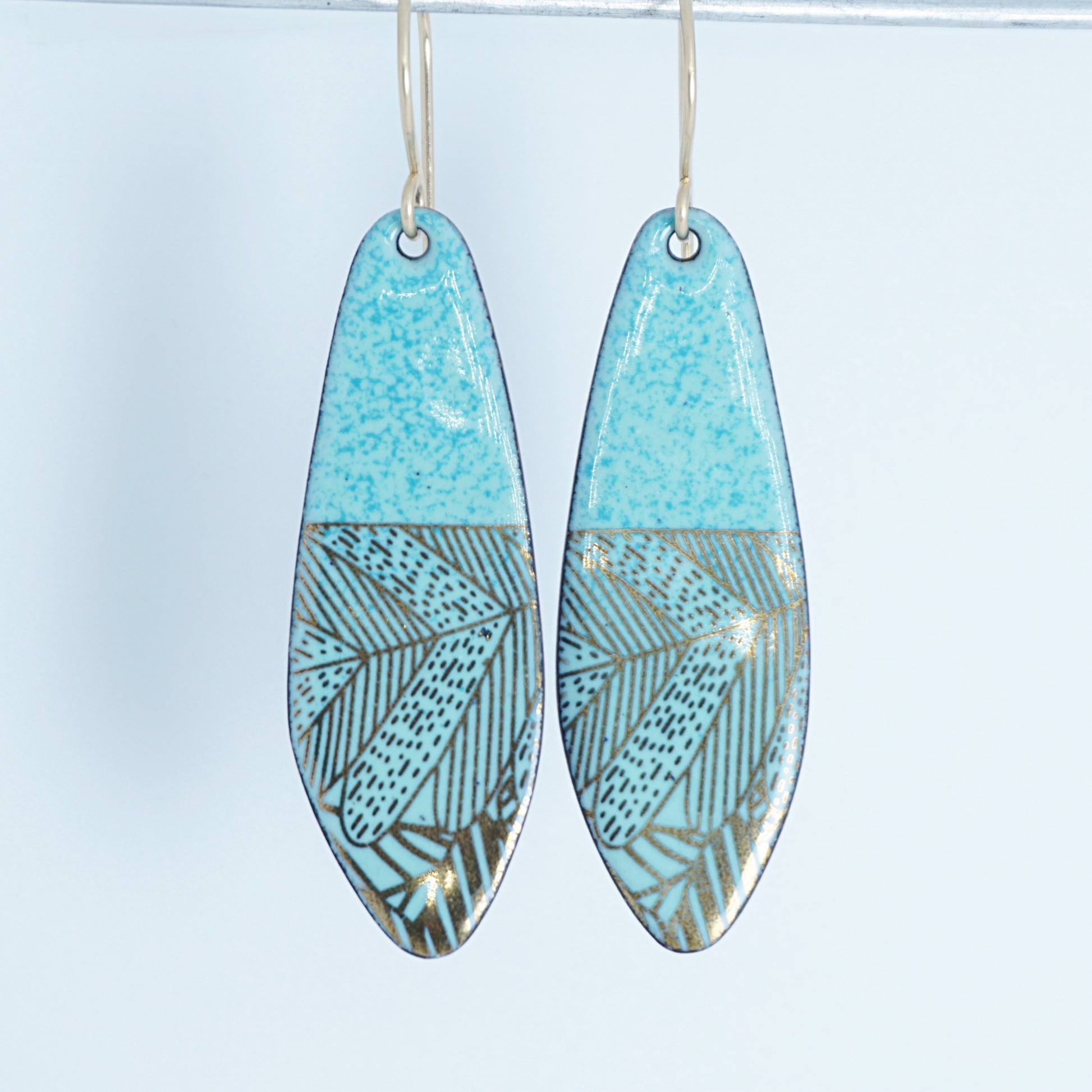 A pair of seafoam green teardrop shaped earrings with gold abstract accents, suspended from thin gold hooks on a white background