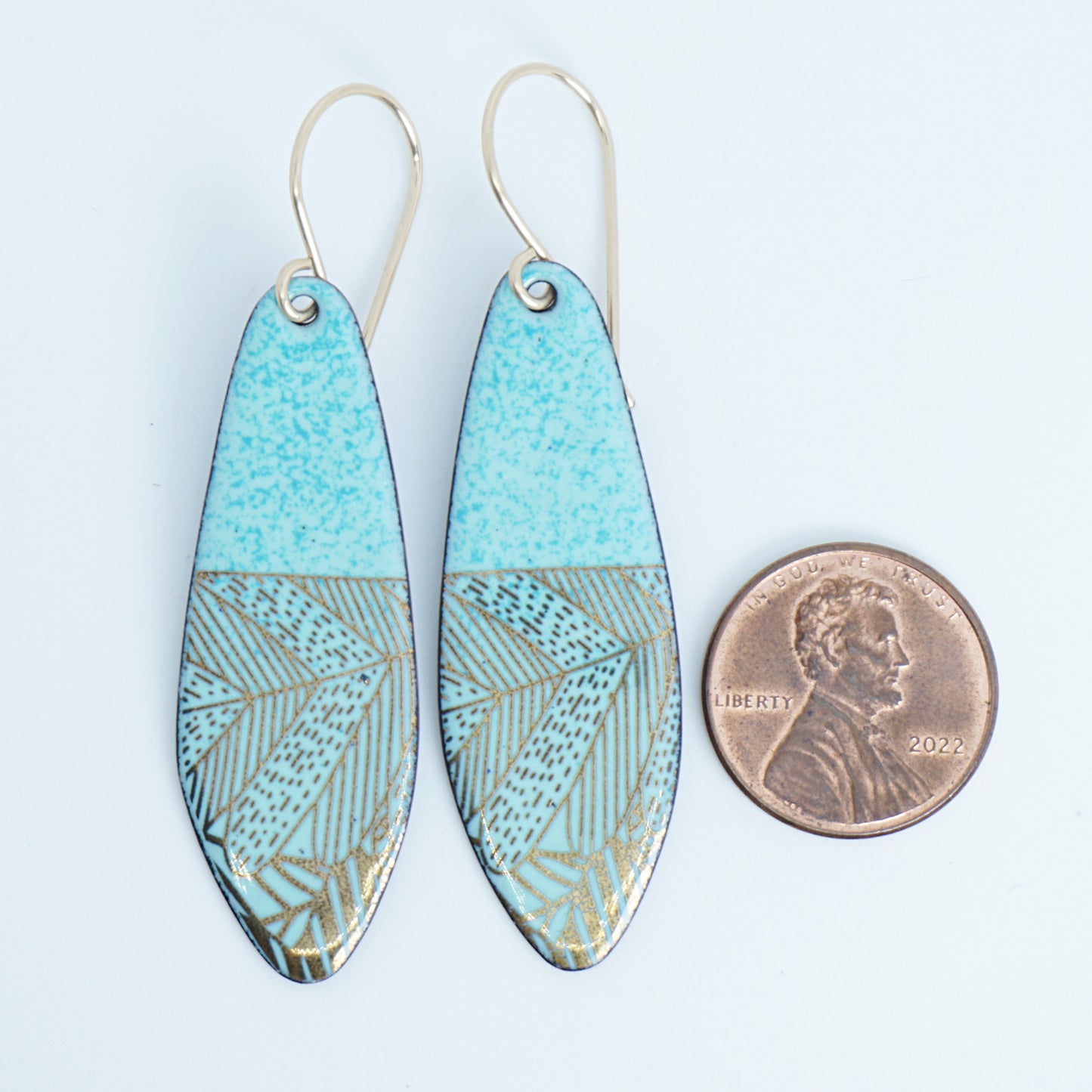 A pair of seafoam green teardrop shaped earrings with gold abstract accents, suspended from thin gold hooks, next to a penny for scale, on a white background