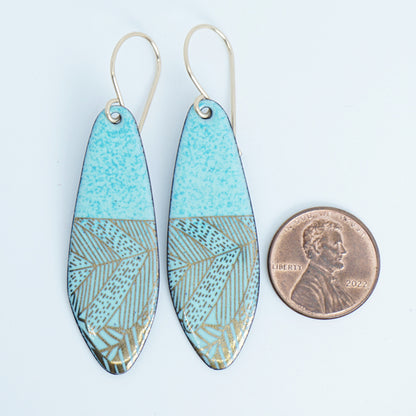 A pair of seafoam green teardrop shaped earrings with gold abstract accents, suspended from thin gold hooks, next to a penny for scale, on a white background