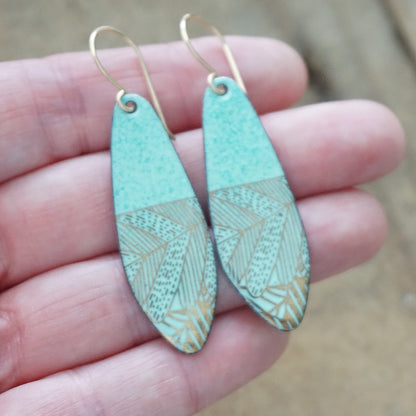 Hand holding A pair of seafoam green teardrop shaped earrings with gold abstract accents, suspended from thin gold hooks on a wooden background