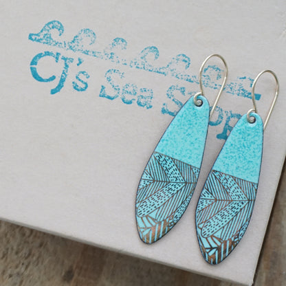 A pair of seafoam green teardrop shaped earrings with gold abstract accents, suspended from thin gold hooks on a gift box