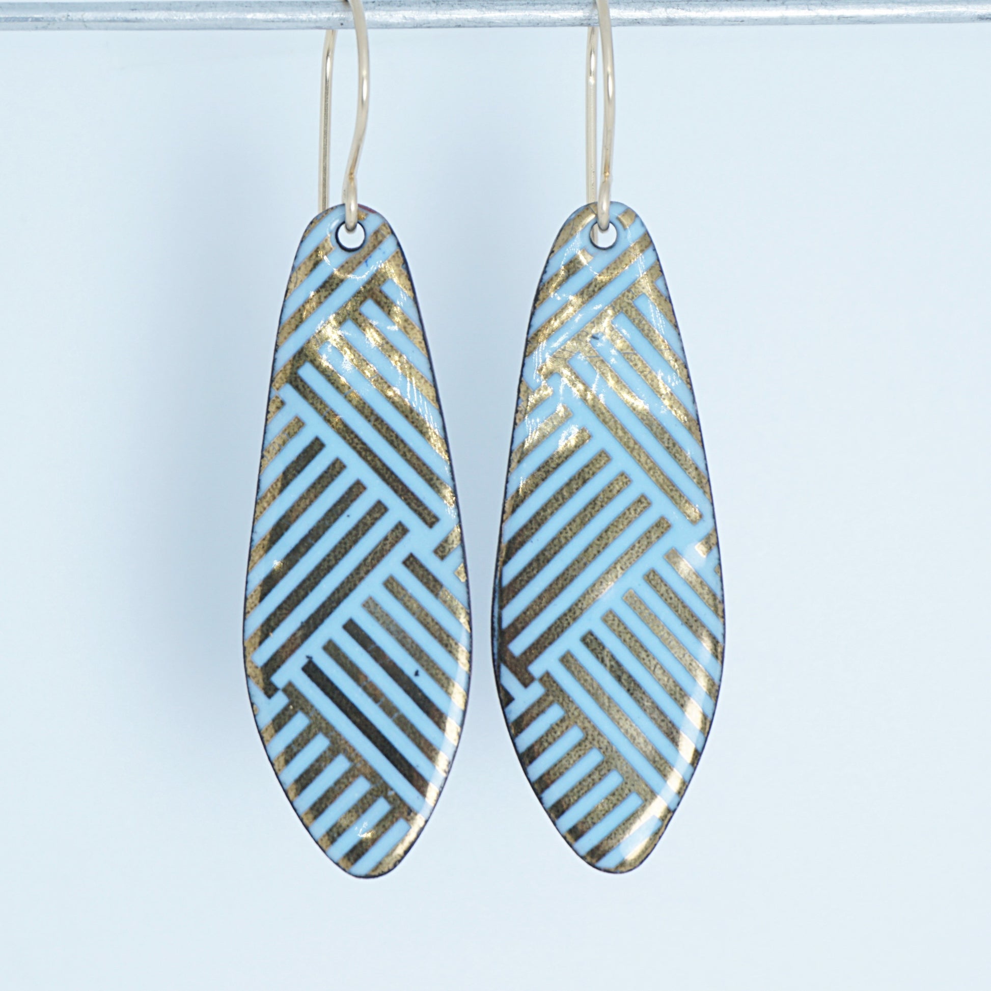 A pair of teardrop-shaped drop earrings with a light blue and gold geometric pattern, hanging from thin gold wire hooks against a white background.