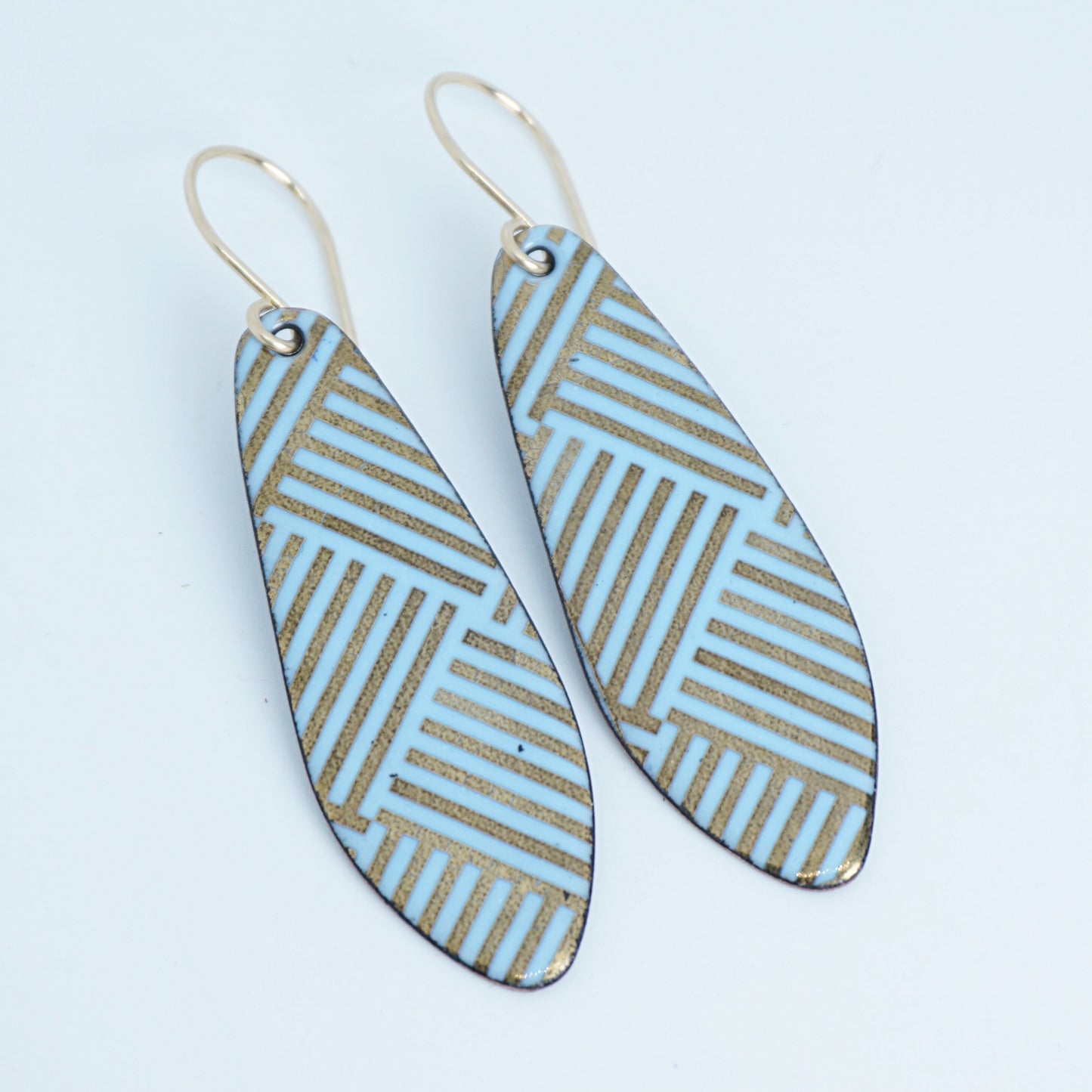 A pair of teardrop-shaped drop earrings with a light blue and gold geometric striped pattern, hanging from thin gold hooks on a white background.