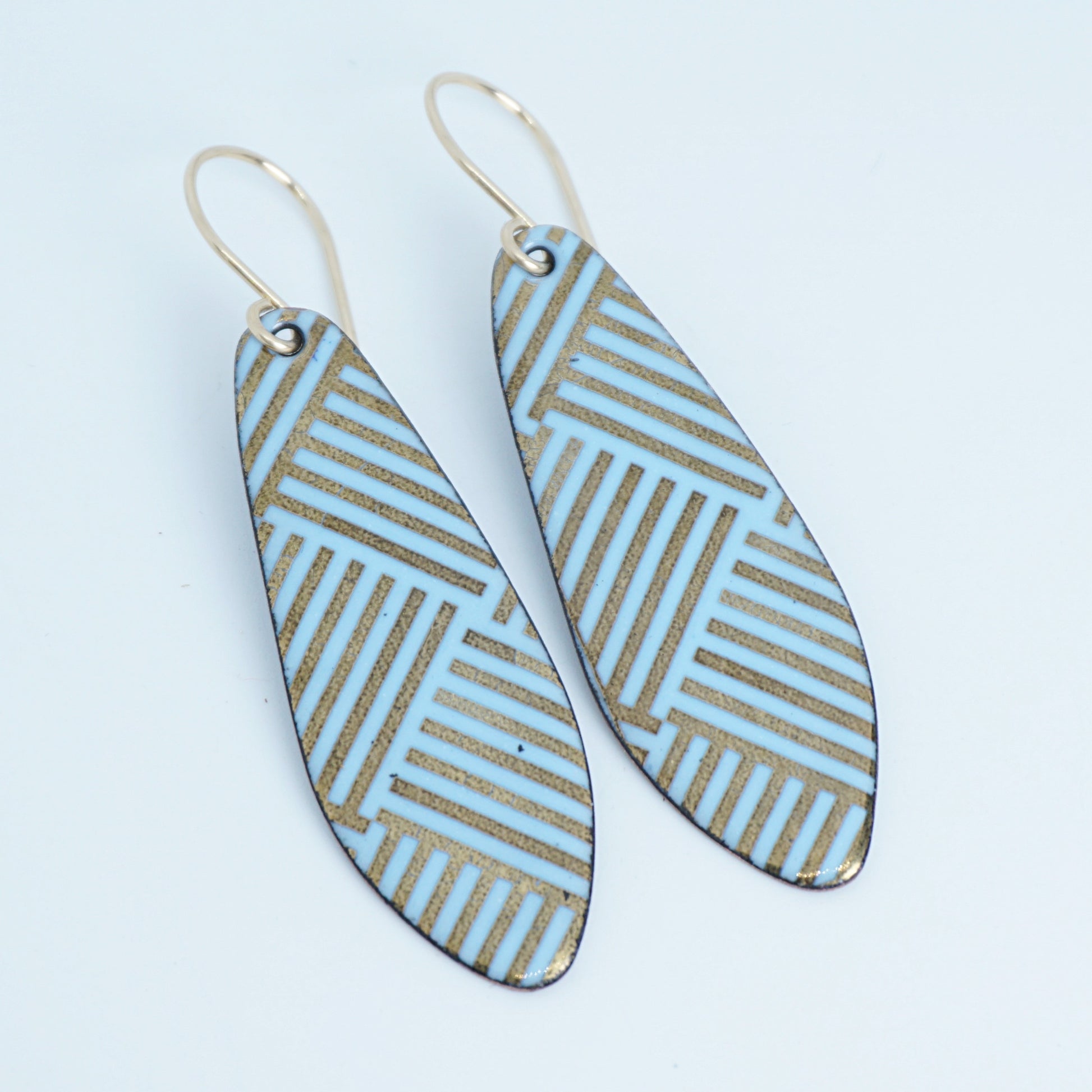 A pair of teardrop-shaped drop earrings with a light blue and gold geometric striped pattern, hanging from thin gold hooks on a white background.
