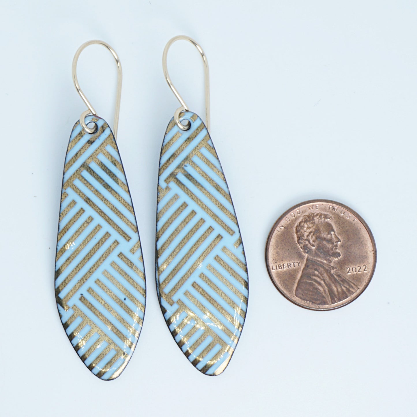 A pair of dangling teardrop earrings with a light blue and gold geometric pattern, shown next to a penny for scale on a white background.