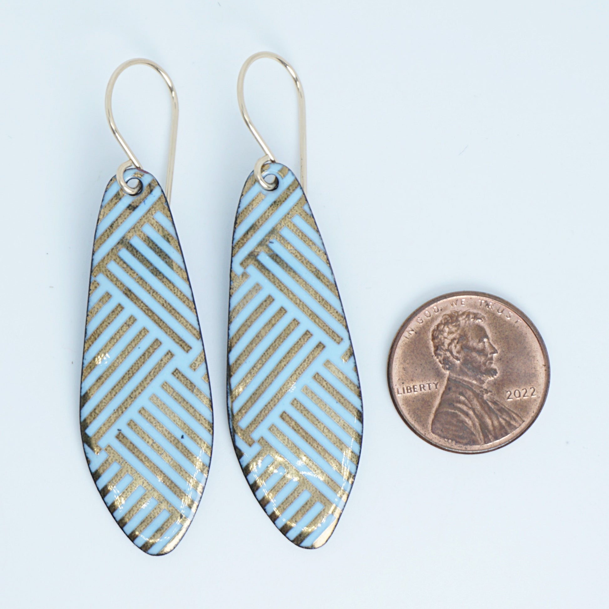 A pair of dangling teardrop earrings with a light blue and gold geometric pattern, shown next to a penny for scale on a white background.