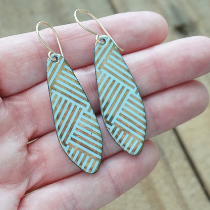 A pair of teardrop-shaped drop earrings with a light blue and gold geometric pattern, held in a person's hand against a blurred wooden background.