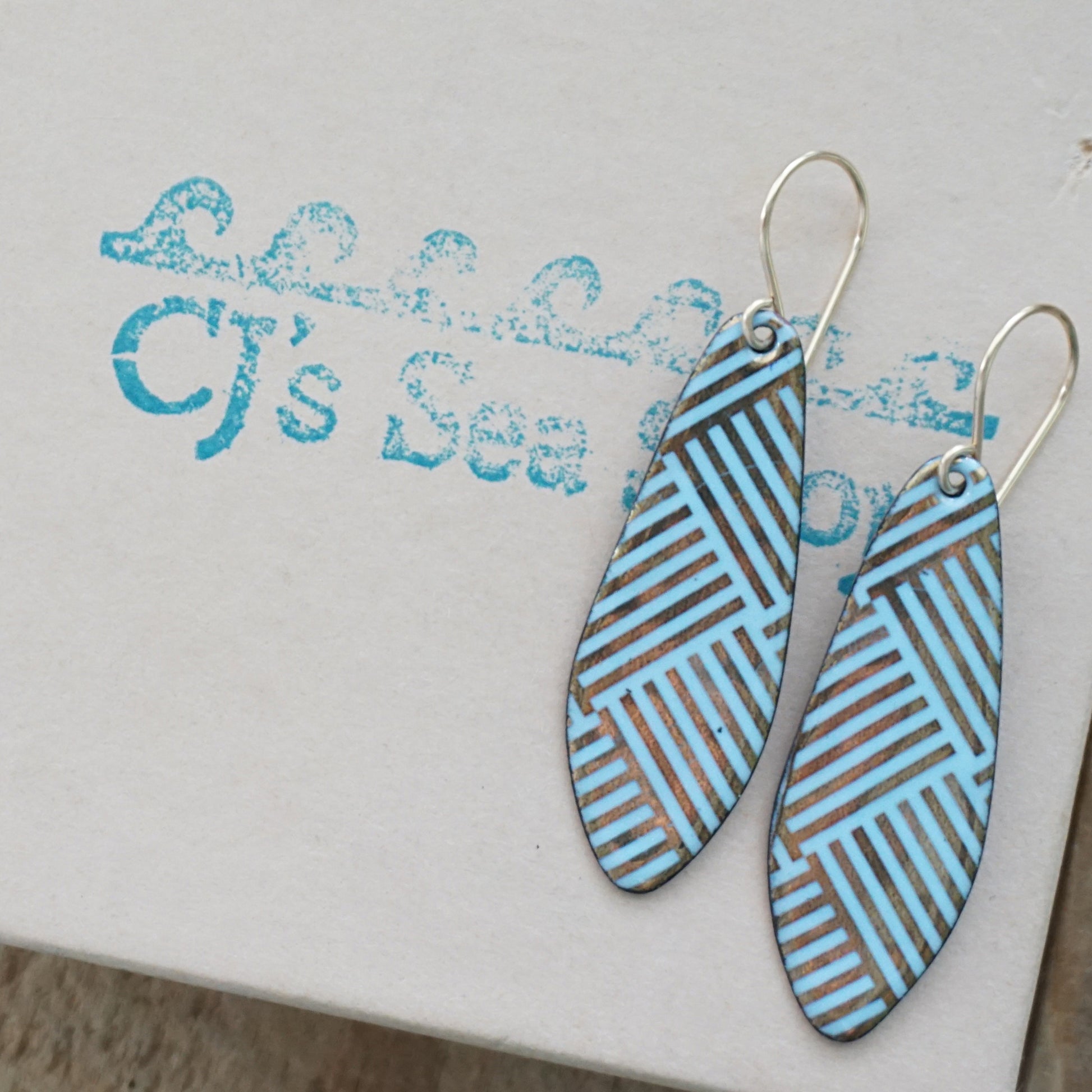 A pair of teardrop-shaped dangle earrings with a light blue and gold geometric striped pattern, hanging from silver fishhook wires on a gift box.